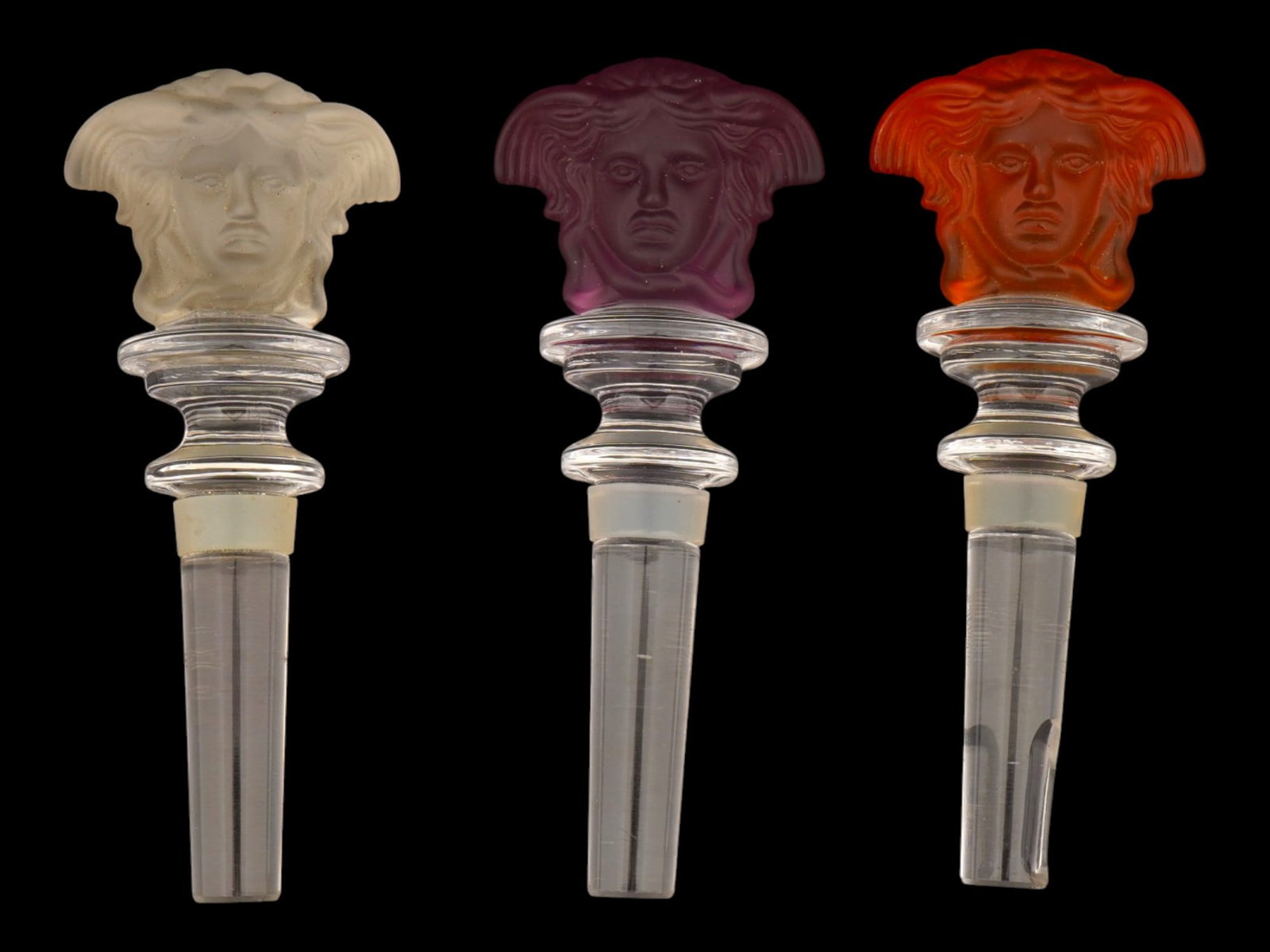 GLASS BOTTLE STOPPERS PRODUCED BY ROSENTHAL FOR VERSACE: A collection of three glass bottle stoppers produced by Rosenthal for Versace. Each features the signature Medusa head motif in relief. The set includes examples in frosted cream, amethyst, and amber 
