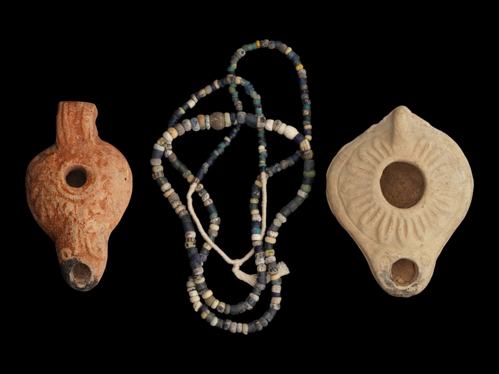 ANCIENT ROMAN AND EGYPTIAN JEWELRY AND OIL LAMPS: A lot of three Ancient Roman artifacts. Imperial Rome era, 100 to 300 AD. A glass beaded necklace. A pair of terracotta oil lamps adorned with ornaments. The lot also includes an ancient Egyptian mosa