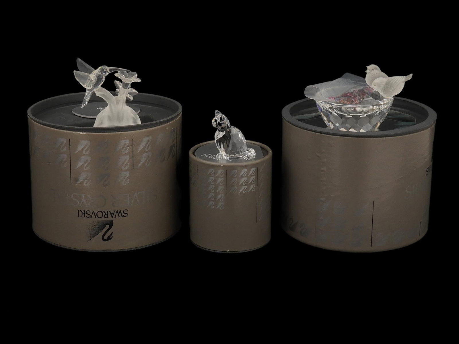 GROUP OF SWAROVSKI CRYSTAL FIGURINES IN ORIGINAL BOXES: A group of Swarovski Crystal figurines, presented in their original gray cylindrical boxes used from the block SC logo period through the adoption of the swan logo in 1989, including a two-hummingbird