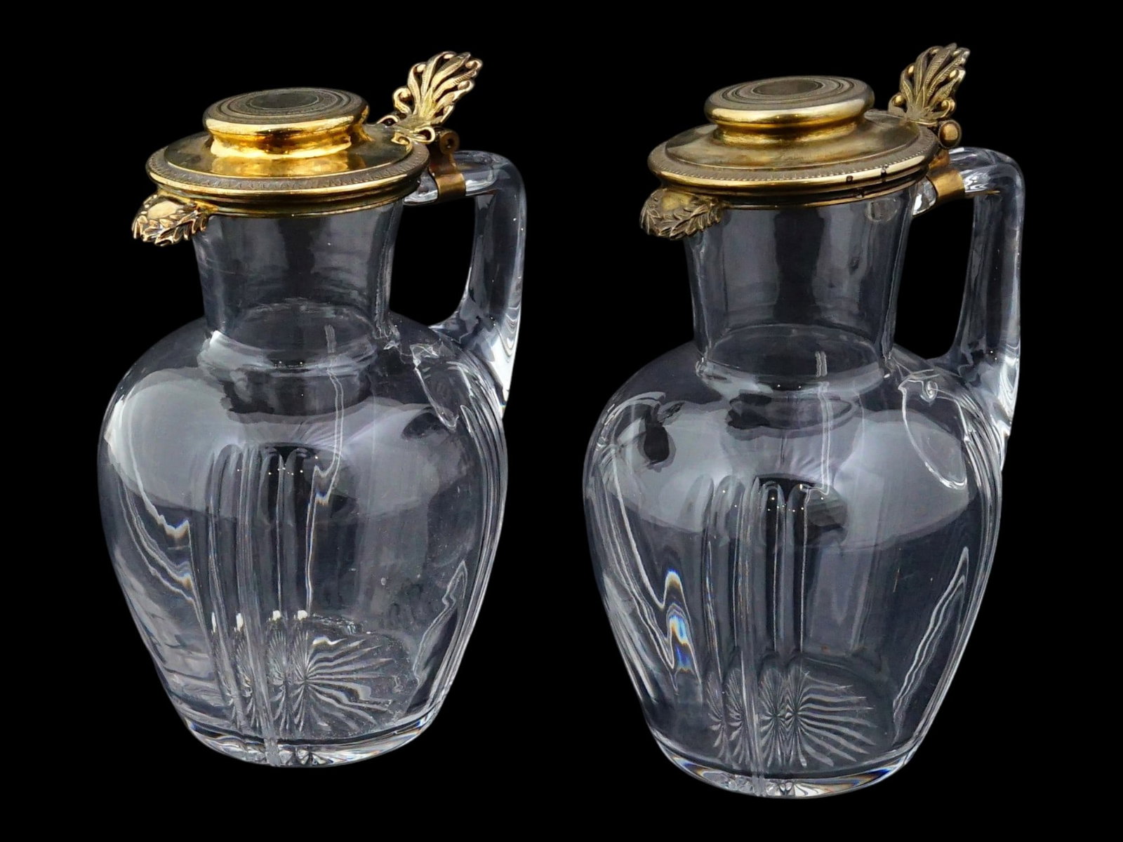ANTIQUE FRENCH CRYSTAL SYRUP JUGS W GILT SILVER LIDS: A pair of antique French crystal syrup jugs with gilt silver lids. The jugs are made of high-quality clear crystal with a faceted relief pattern on the bodies. The gilt silver lids are decorated with 