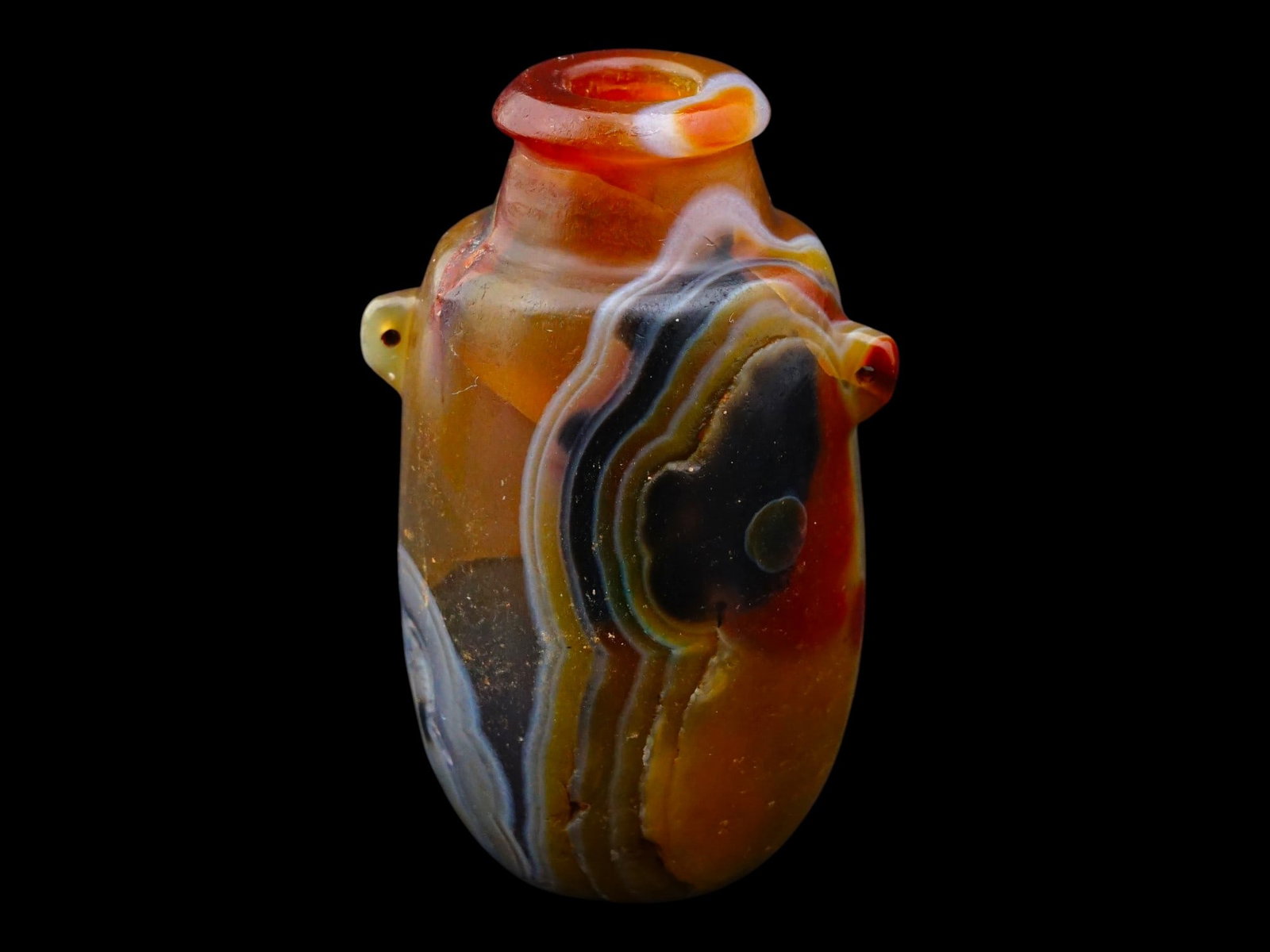 ROMAN BANDED AGATE AMPHORISKOS MINIATURE VESSEL: A Roman carved miniature vessel, or amphoriskos, is fashioned from a single block of translucent banded agate, displaying a rich palette of carnelian, honey, and charcoal tones. The piece features an 