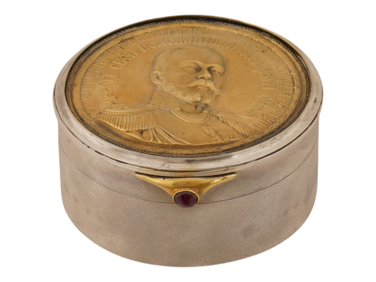 RUSSIAN SILVER AND RUBY TRINKET BOX NICHOLAS II: A Russian silver trinket box. Round piece, the clasp is set with a ruby cabochon. The gilt hinged lid bears a relief portrait of the Emperor Nicholas II and a quote from the Gospels. Gold wash interio