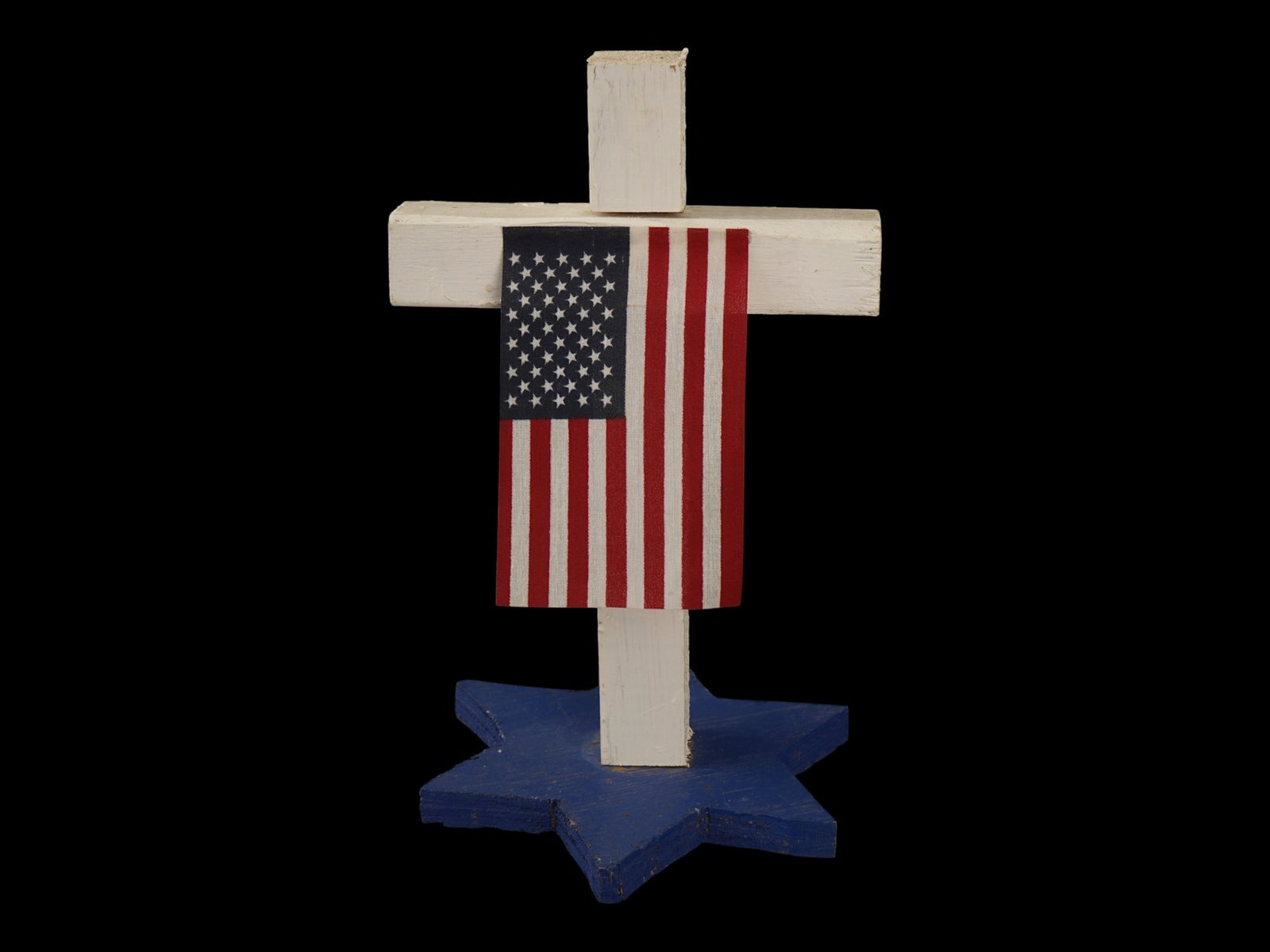 VINTAGE HANDMADE AMERICAN FLAG PANEL FOLK ART PIECE: A vintage handmade American folk art piece featuring a white wooden cross decorated with an American flag panel. The cross is mounted on a blue multi-pointed star base, suggesting a commemorative mili