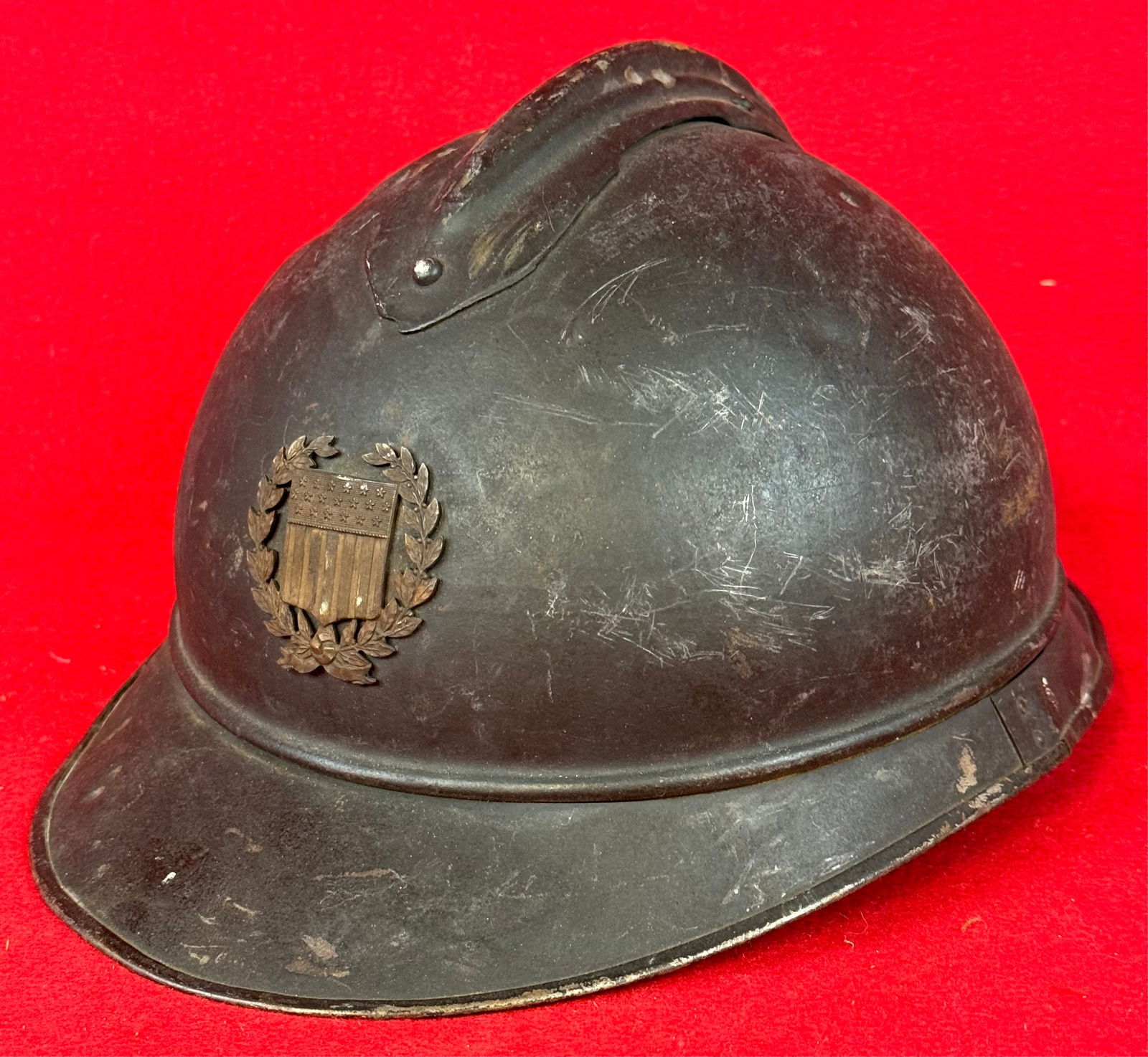 French Model 26 "Adrian" Helmet With Battle Damage: French Model 26 "Adrian" combat helmet with brass or bronze French Republic shield on front and dark gray finish. Helmet exhibits battle damage . Helmet lacks liner but does retain its chinstrap.