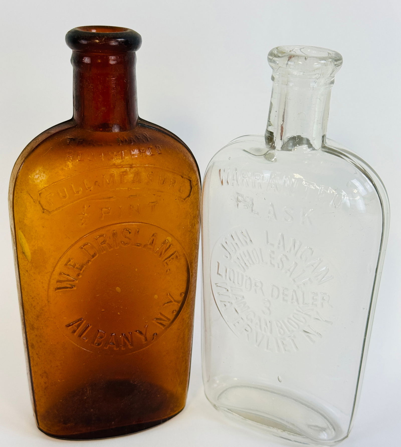 Lot 2 Strap Sided Flasks Drislane Albany / Langan Watervliet: Lot of 2 strap sided whiskeys each being a pint capacity with the first amber with a round slug plate at front embossed W E Drislane Albany N.Y. Bottle has some wear and exterior stain but is free of