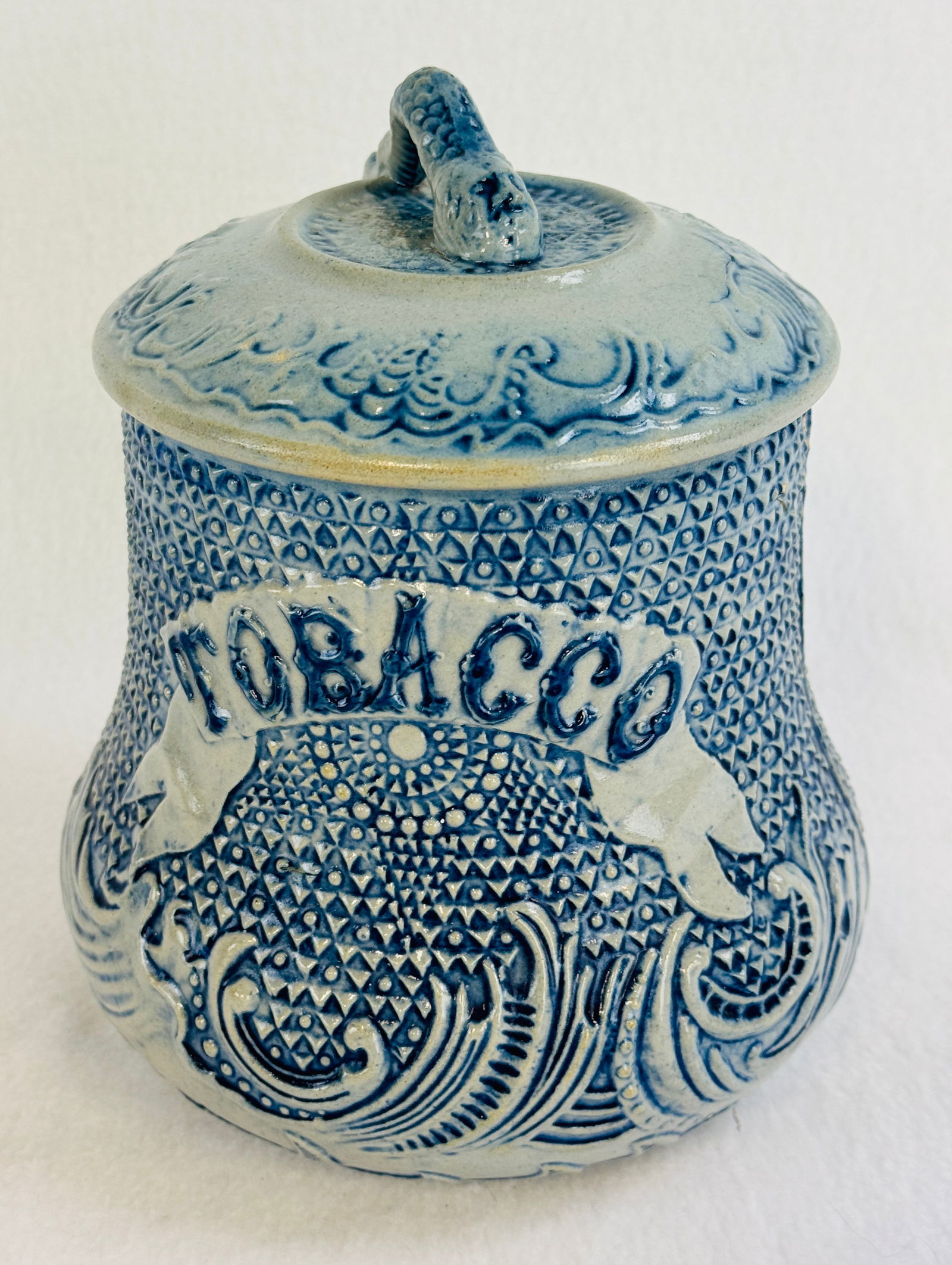 Blue & White Molded Stoneware Tobacco Jar: 7 1/2" blue and white molded stoneware tobacco jar with lid which is embossed TOBACCO at front. Finial on lid has the face of a man molded into it. Condition - lid is excellent, body of jar is cracked