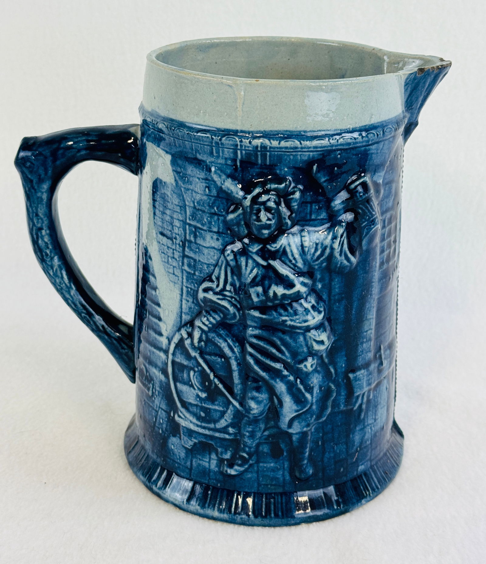 8 1/2" Blue & White Molded Stoneware Pitcher: 8 1/2" blue and white molded stoneware pitcher which has scenes of a man and woman holding up steins etc she in a interior home setting he in a wine cellar. Condition - several small chips to spout.