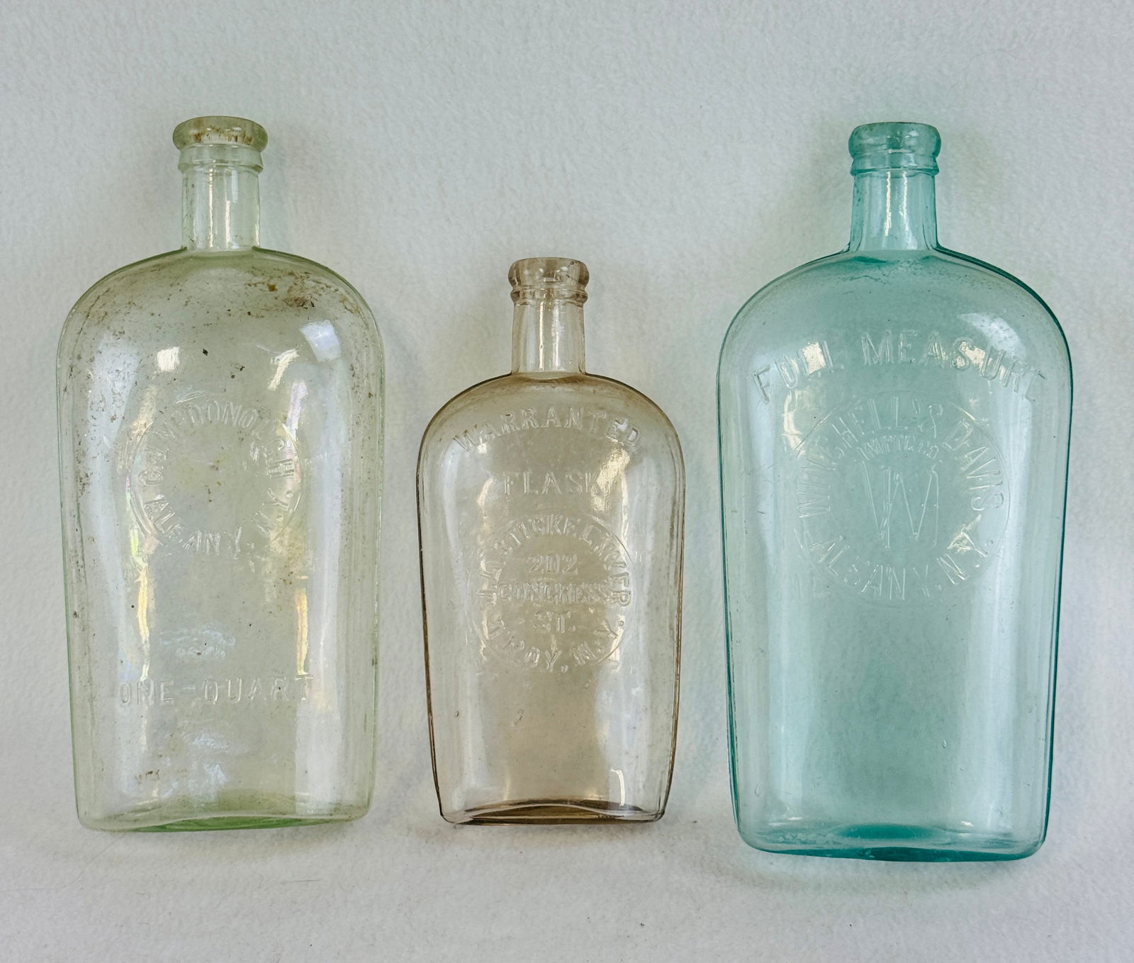 Lot 3 Strap Sided Flasks: Lot of three strap sided whiskey flasks each with a slug plate front. The first is in a very light green quart embossed C. J. McDonough Albany N.Y.; another is a aquamarine quart embossed in a round f