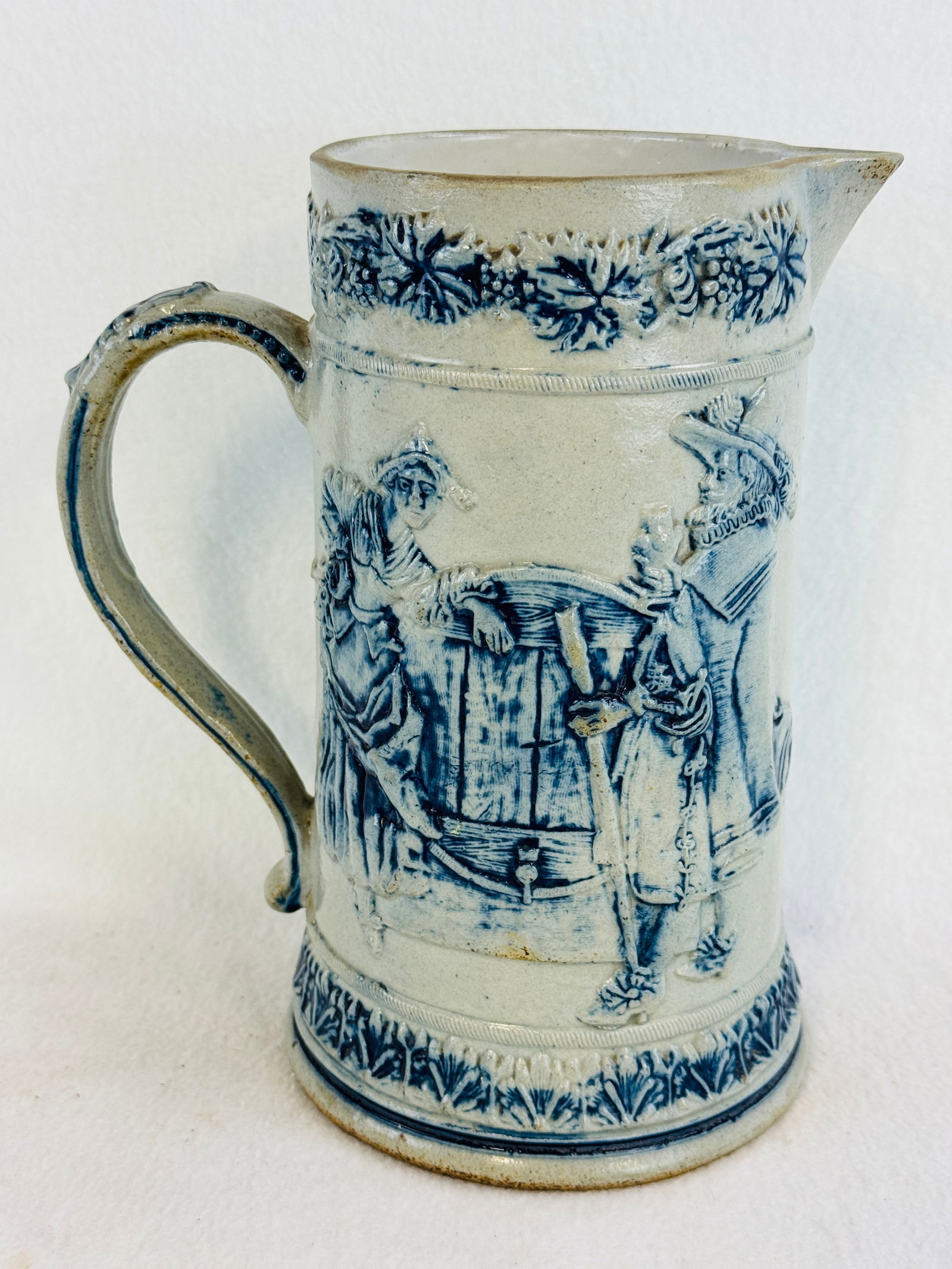 Blue & White Molded Stoneware Pitcher: 9 1/4" blue and white molded stoneware pitcher which has sides decorated with two designs one shows three adults and a boy drinking from stein or mug while the other has a couple in early attire in a
