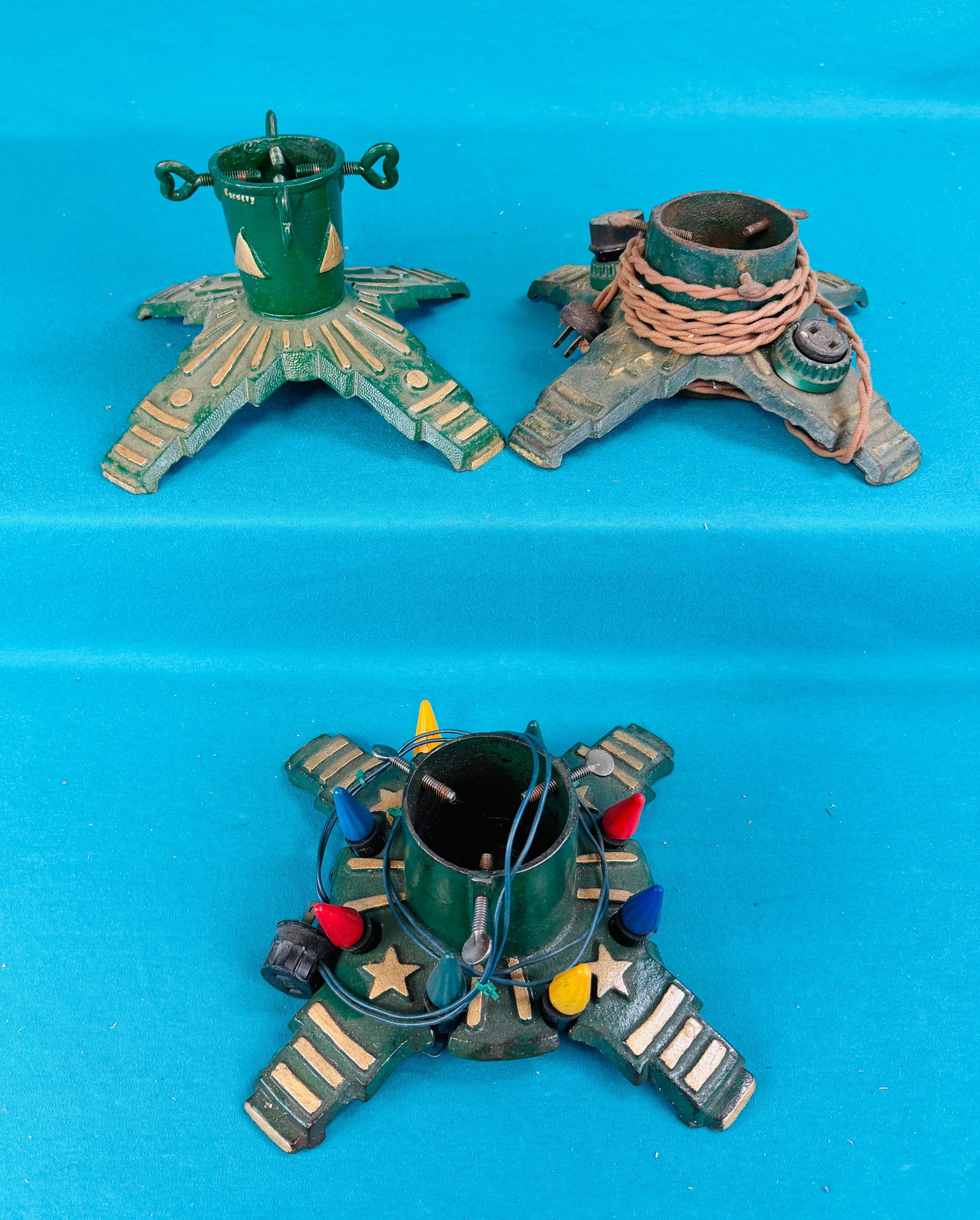 3 Vtg Cast Iron Christmas Tree Stands 1 Lighted: Lot of three different variants of cast iron Christmas Tree stands one of which is lighted with period bulbs. Two of the stands are cast with a star motif as shown. Condition appears to retain their o