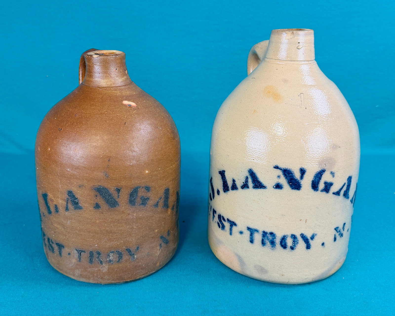 Lot 2 19th C Stoneware Advertising Jugs: Lot of two 19th century one gallon capacity straight sided stoneware jugs from West Troy (Watervliet) New York. The first jug is stenciled J Langan West Troy N.Y. and aside from a small chip on the sp