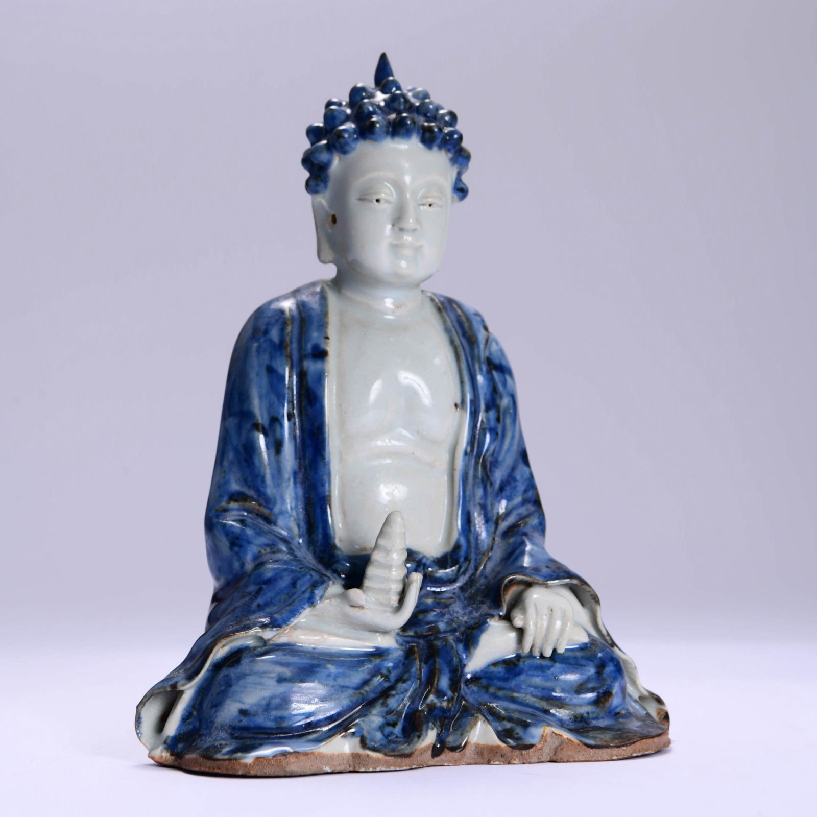 PORCELAIN BLUE & WHITE SAKYAMUNI STATUE: The figure is cast seated in dhyanasana. He is depicted wearing a robe that exposes his chest and is decorated with incised floral hems. His face is modelled with a serene expression which is framed l