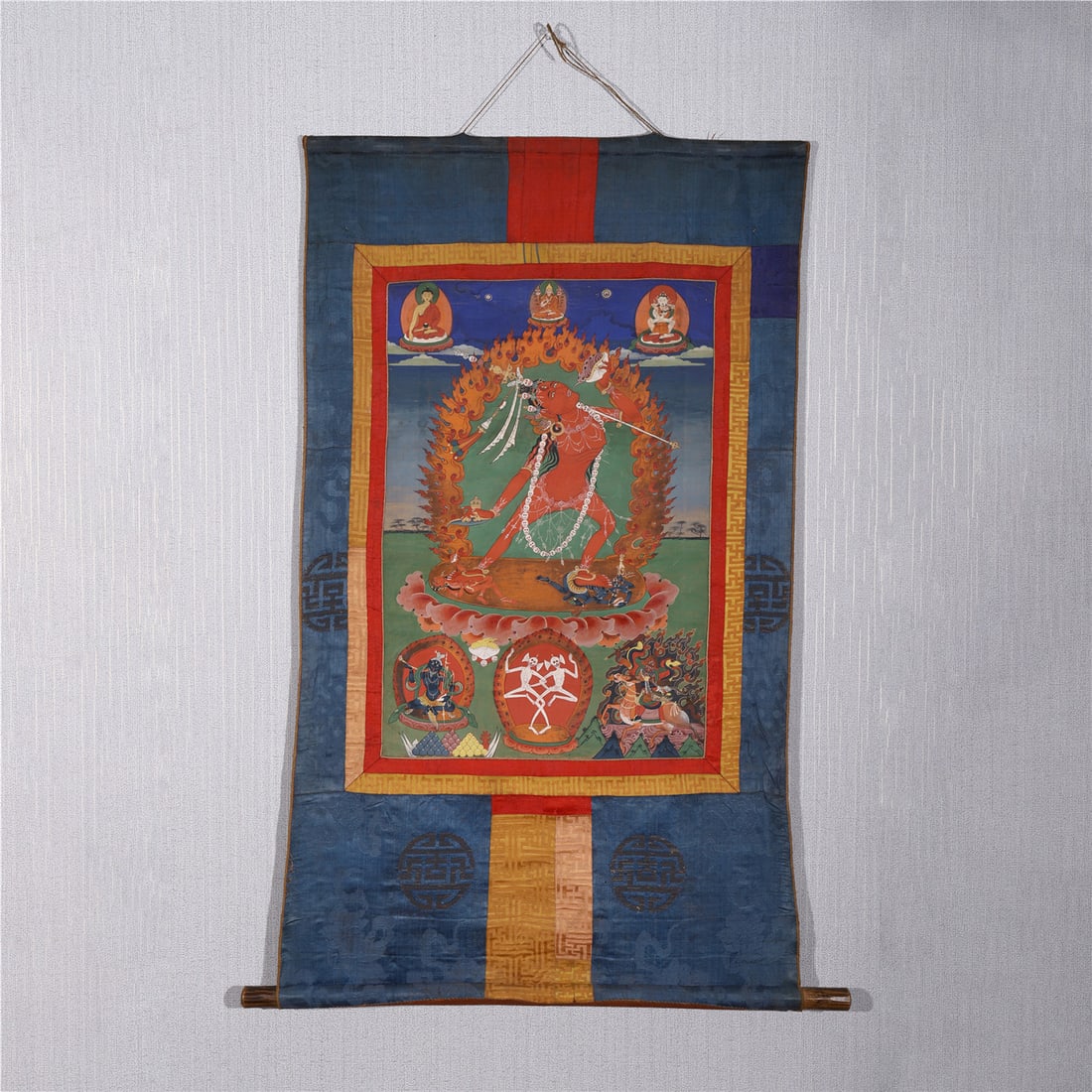 CHINESE TIBETAN THANGKA WRATHFUL DEITY: Dimensions: L. 83 cm. W. 50 cm. This vibrant Chinese Tibetan thangka depicts a wrathful Buddhist deity in vivid red tones, standing within a flaming mandorla against a blue sky and green landscape, fl