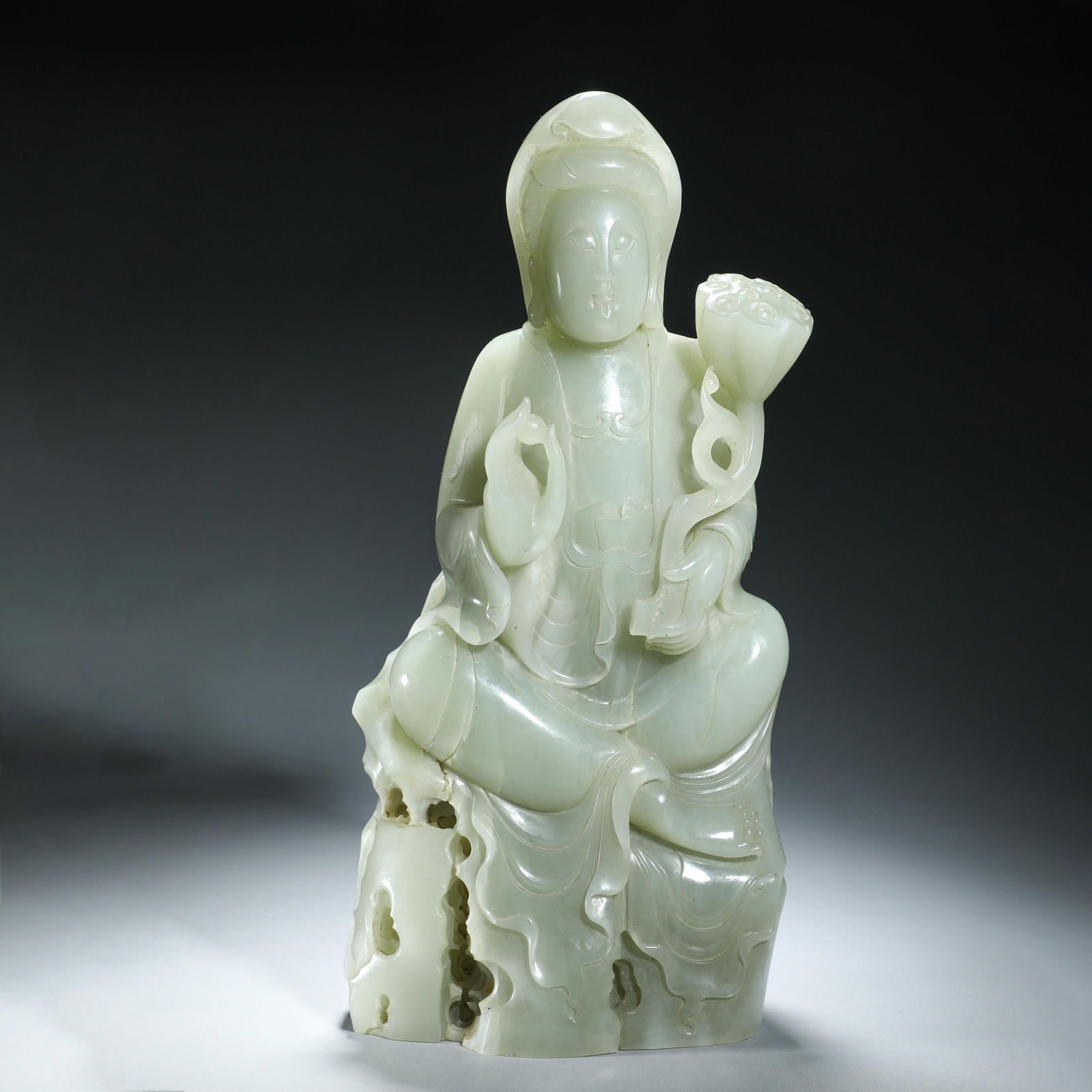 CHINESE HETIAN JADE GUANYIN STATUE: Dimensions: L. 15 cm. H. 32 cm. Weight 4500 grams. This Chinese Hetian jade carving depicts Guanyin seated on a rocky base, holding a lotus bud with a serene expression, measuring 15 cm in length, 32