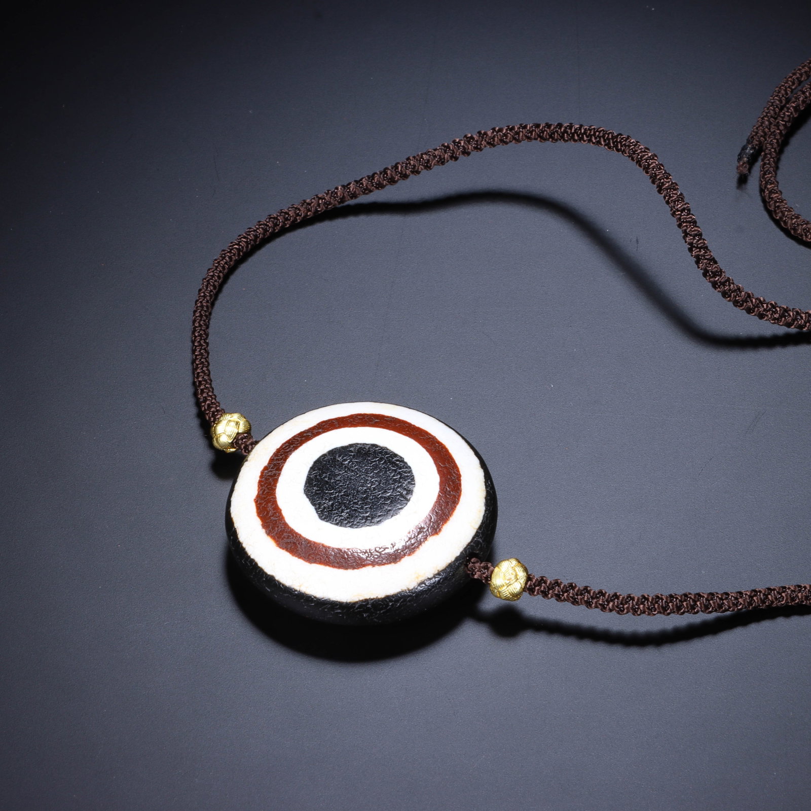 A CHINESE EYE DZI BEAD PENDANT NECKLACE: Dimensions: L. 3.8 cm. This elegant Chinese necklace is centered ? a striking disc-shaped agate dzi bead, featuring a bold concentric "eye" pattern with layers of black, reddish-brown, and creamy whit