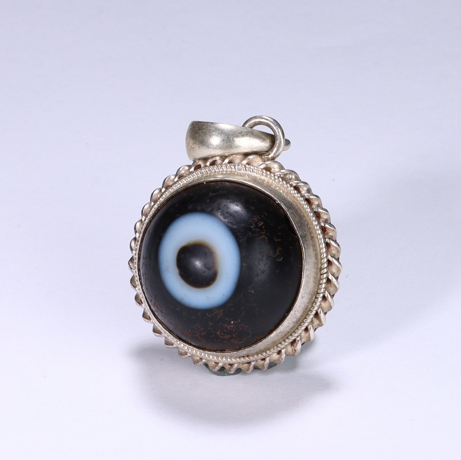 A CHINESE SILVER-MOUNTED EYE DZI BEAD PENDANT: Dimensions: L. 2.5 cm. This exceptional Chinese pendant centers on a disc-shaped agate dzi bead, showcasing a striking concentric "eye" motif with layers of deep black, bright white, and warm brown. T