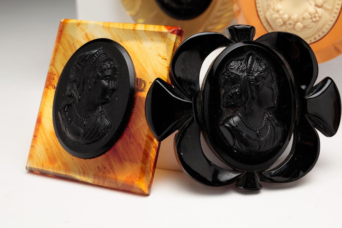SEVEN BAKELITE & CELLULOID CAMEOS.: American, 2nd Qtr - 20th century. Group includes: one black oval pendant with celluloid oval links; one black rectangular pendant with cream color applied cameo, leather neck strap; three booches of v