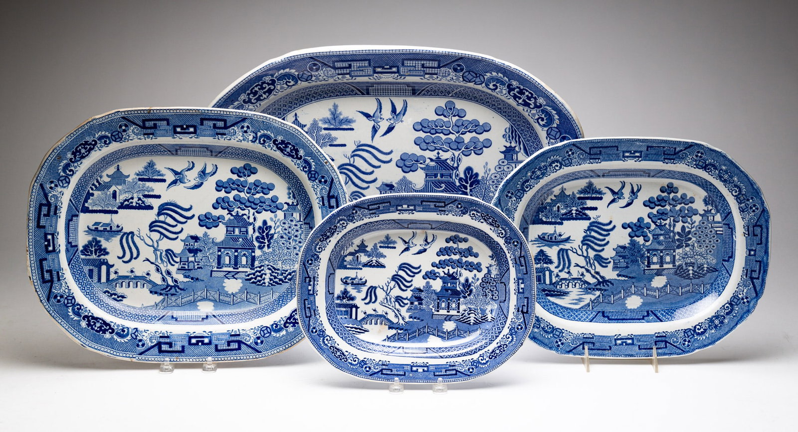 FOUR STAFFORDSHIRE "BLUE WILLOW" PLATTERS.: England, Second Quarter 19th Century. All four have combed backs. Smallest has transfer mark on reverse "Warrented, Stafford, Stone China, No. 14". Wear and light small stains. 12.25"w. 9.75"d., next 
