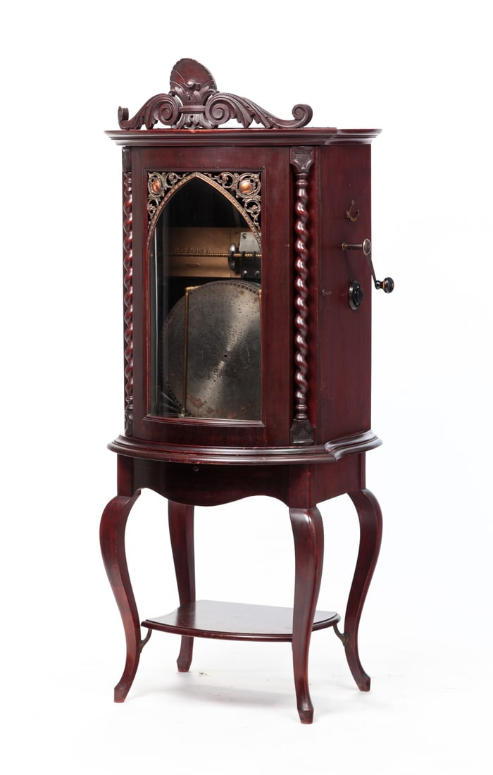 AMERICAN REGINA "STYLE 36" CABINET DISC PLAYER.: Circa 1900. Mahogany. Anthemion and leaf bracket carved cornice, molded cornice, curved glass door with openwork cast nickel plated valance flanked by briar twist columns, lower drawer and shelf and c