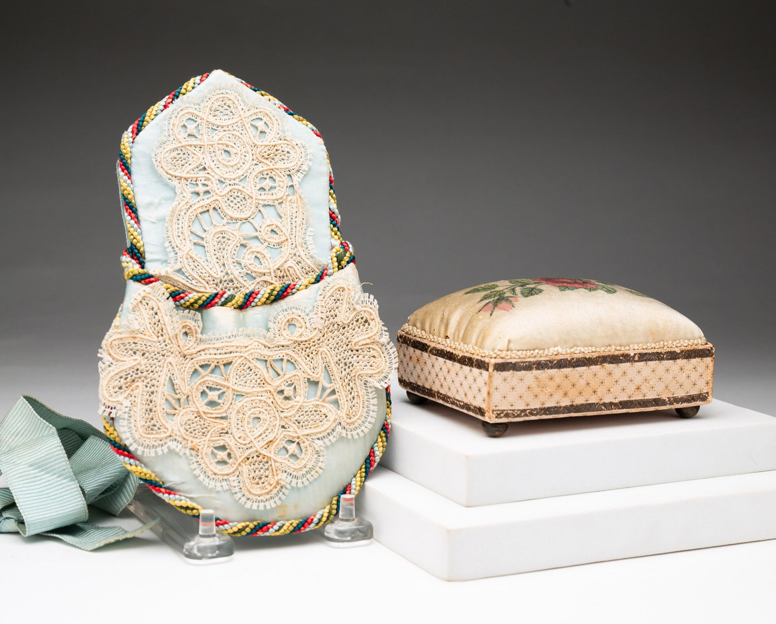 TWO PIECES OF SEWING ACCOUTREMENT.: American, second half 19th Century. A blue satin pocket with lace adornment, multi colored corded edge and grosgrain ribbon belt. A little wear. 7.5"h. and a rectangular pincushion with painted roses,