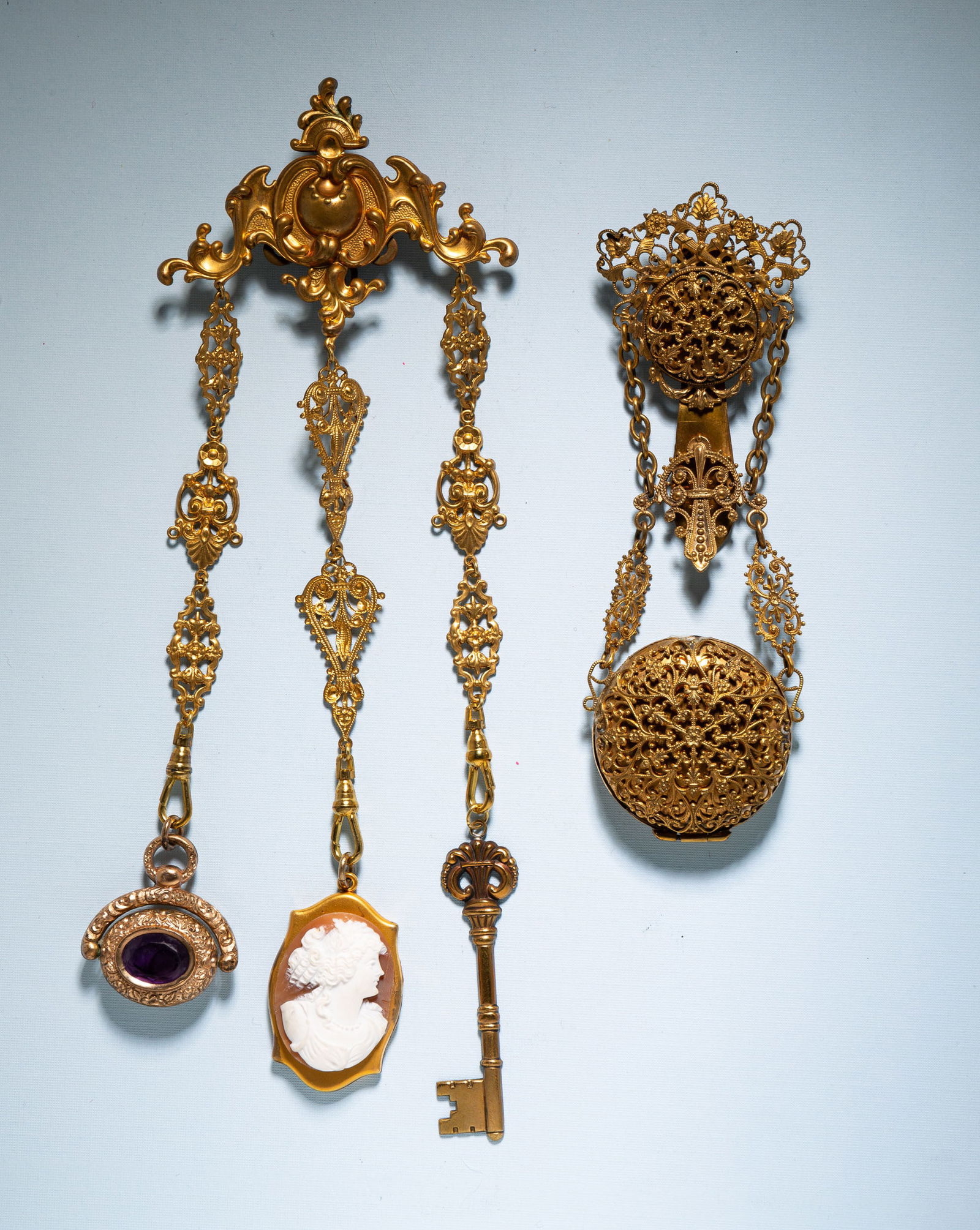 TWO PINCHBECK FILIGREE CHATELAINES.: European, late 19th Century. One with filigree clip and large locket with filigree front and print of classical lady and cherub on the back. 5"l. One with ornate clip and three filigree chains with dr