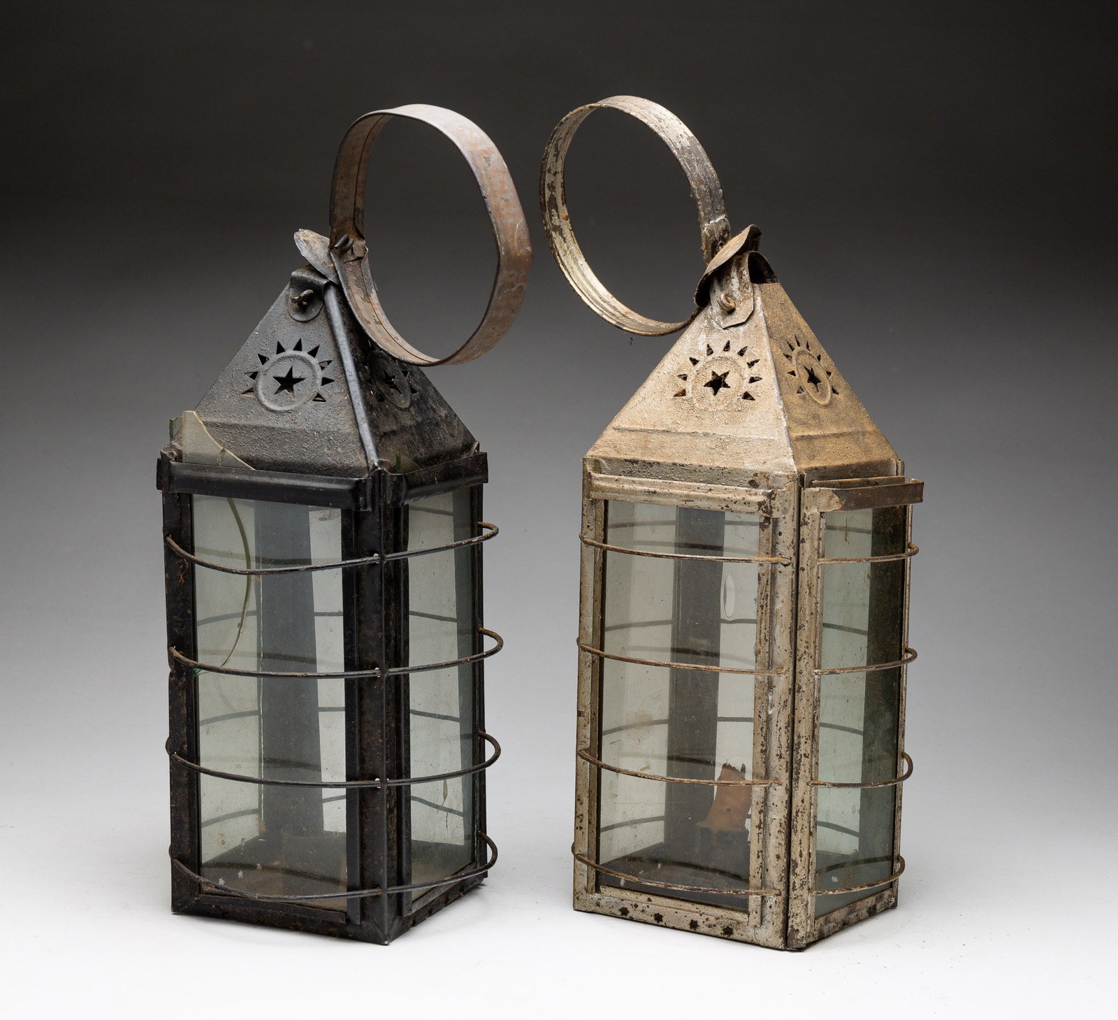 TWO SQUARE PIERCED TIN CANDLE LANTERNS.: American, 19th Century. Two glazed, four-sided tin lanterns with [glass] guards, pyramidal star-pierced tops, and ring handles. With handle, 4.5"w., 16"h. From the collection of the late Audrey Caspar