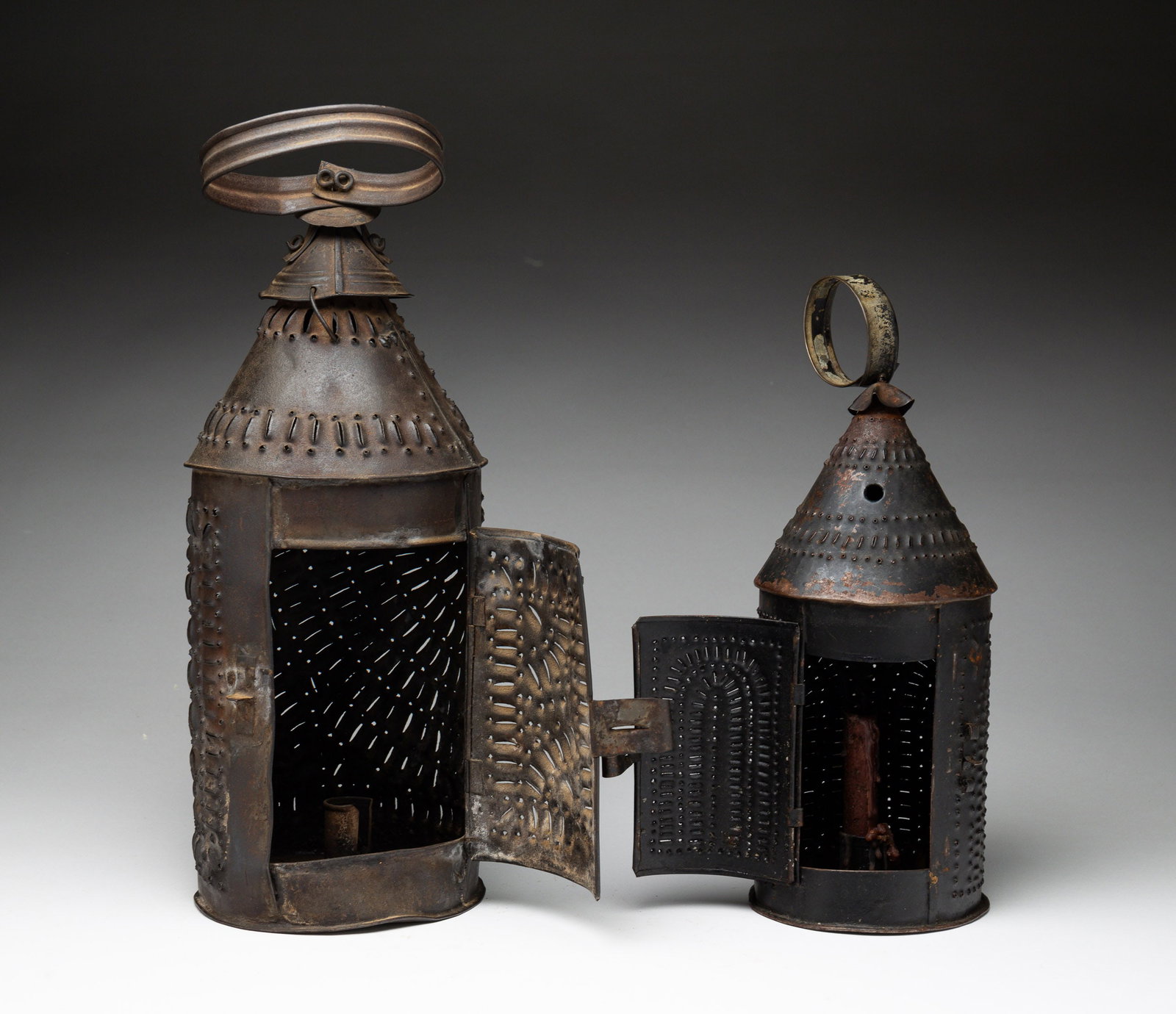 TWO PUNCHED TIN REVERE-TYPE CANDLE LANTERNS.: American, 19th Century. Two cylindrical, punched tin candle lanterns with hinged doors, conical tops, and round hangers. 12"h. and 18.5"h. From the collection of the late Audrey Caspari (New Jersey).