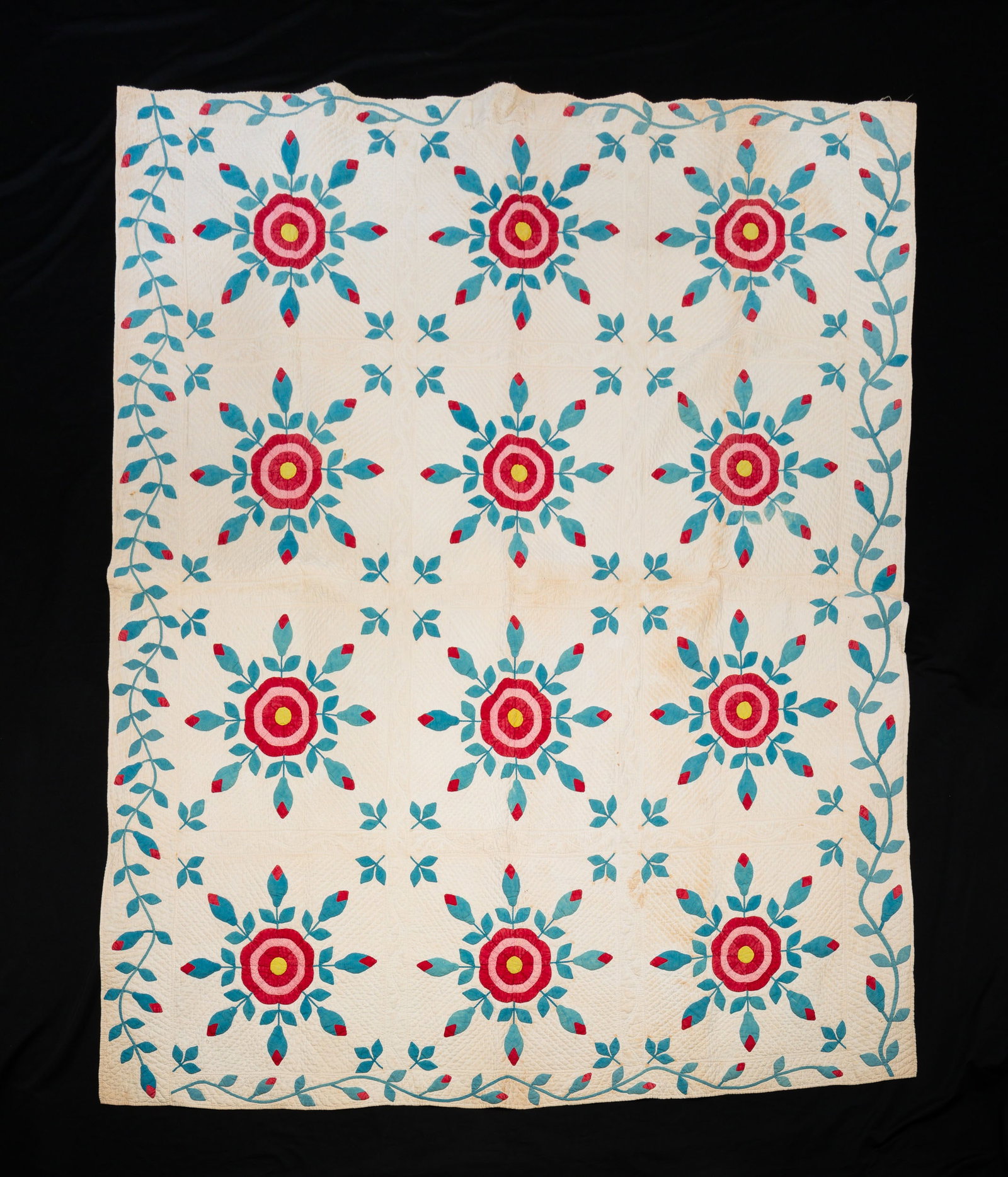 APPLIQUE AND STUFFED QUILT.: American, mid 19th Century. Hand-stitched cotton with twelve pattern blocks surrounded by a floral vine border to all four sides in pieced red, blue, and yellow fabrics. Quilted and vining trapunto wo