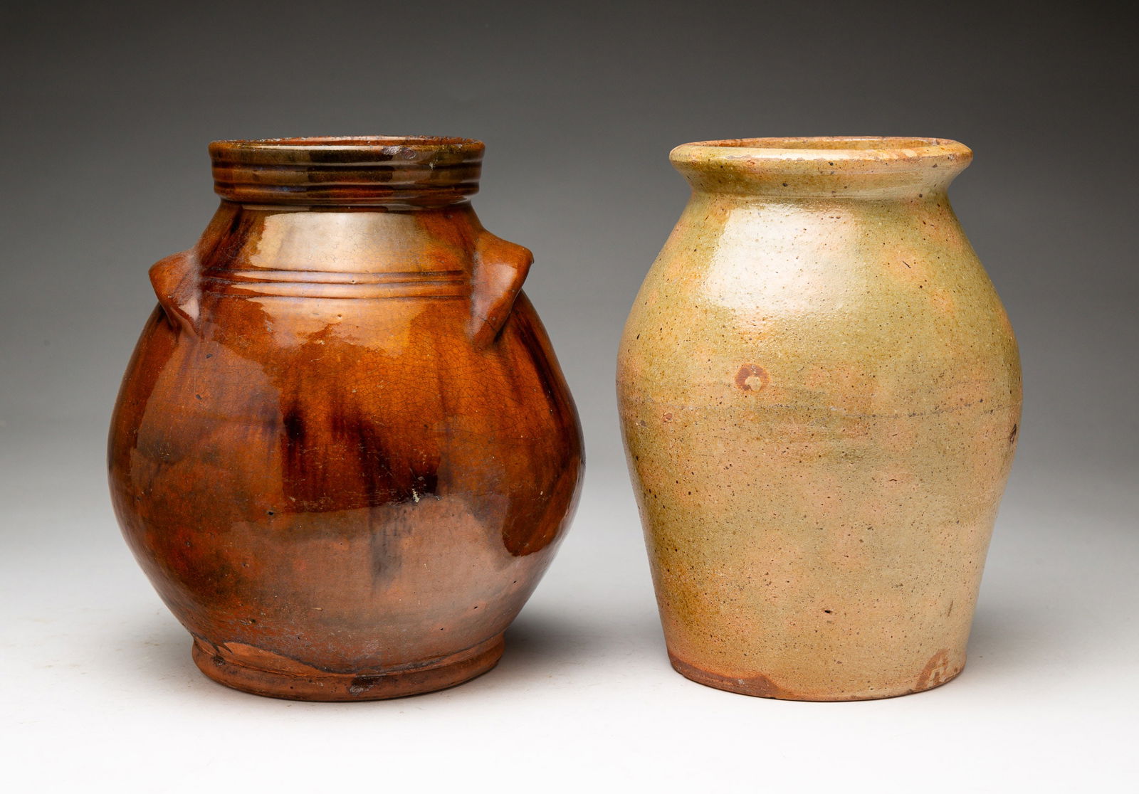 TWO OVOID REDWARE JARS.: American, second half 19th Century. One has a flared lip and tan and green mottled glaze. Wear and glaze flakes. 8.5"h. The second has handles and mottled brown glaze. Glaze flakes and small chips.