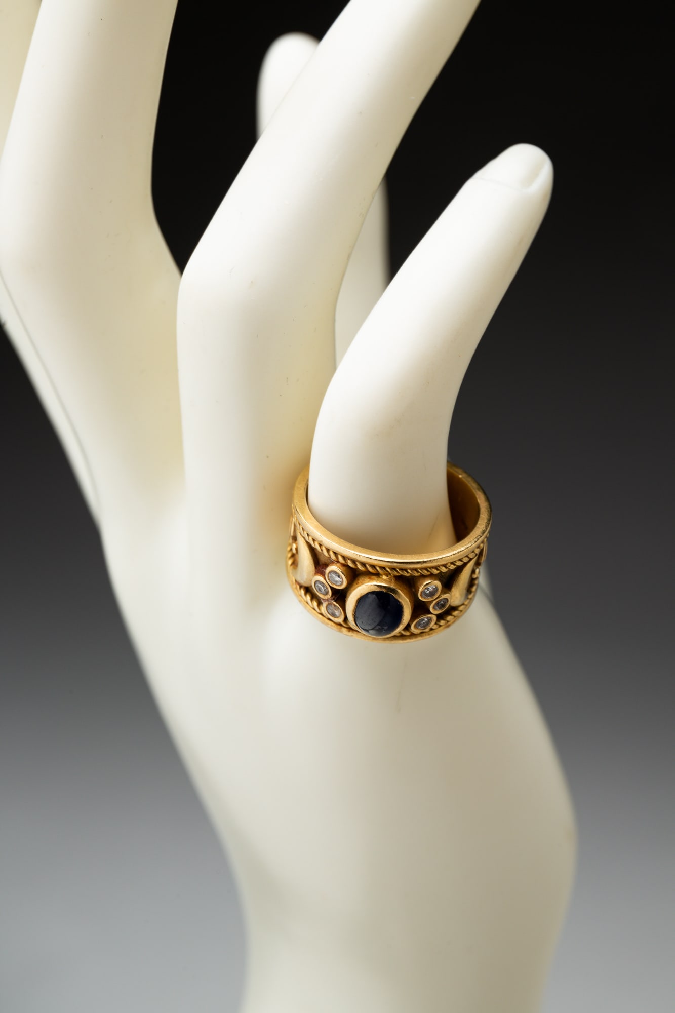 18 KARAT GOLD BAND WITH CABOCHON SAPPHIRE.: American, late 20th Century. Tapered yellow gold band with center bezel set cabochon sapphire and six small accent diamonds. Raised gold design around band. Measures approx.11.5mm at front, ring size 
