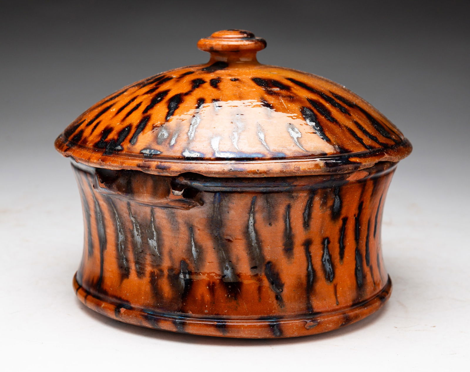LIDDED REDWARE BUTTER CROCK.: American, mid-19th Century. Manganese decoration. Sloped sides, slight footed base and small lug handles. Chips, loss and edge wear, wear and loss to handles and lid, burst pot bubble to bottom, firin