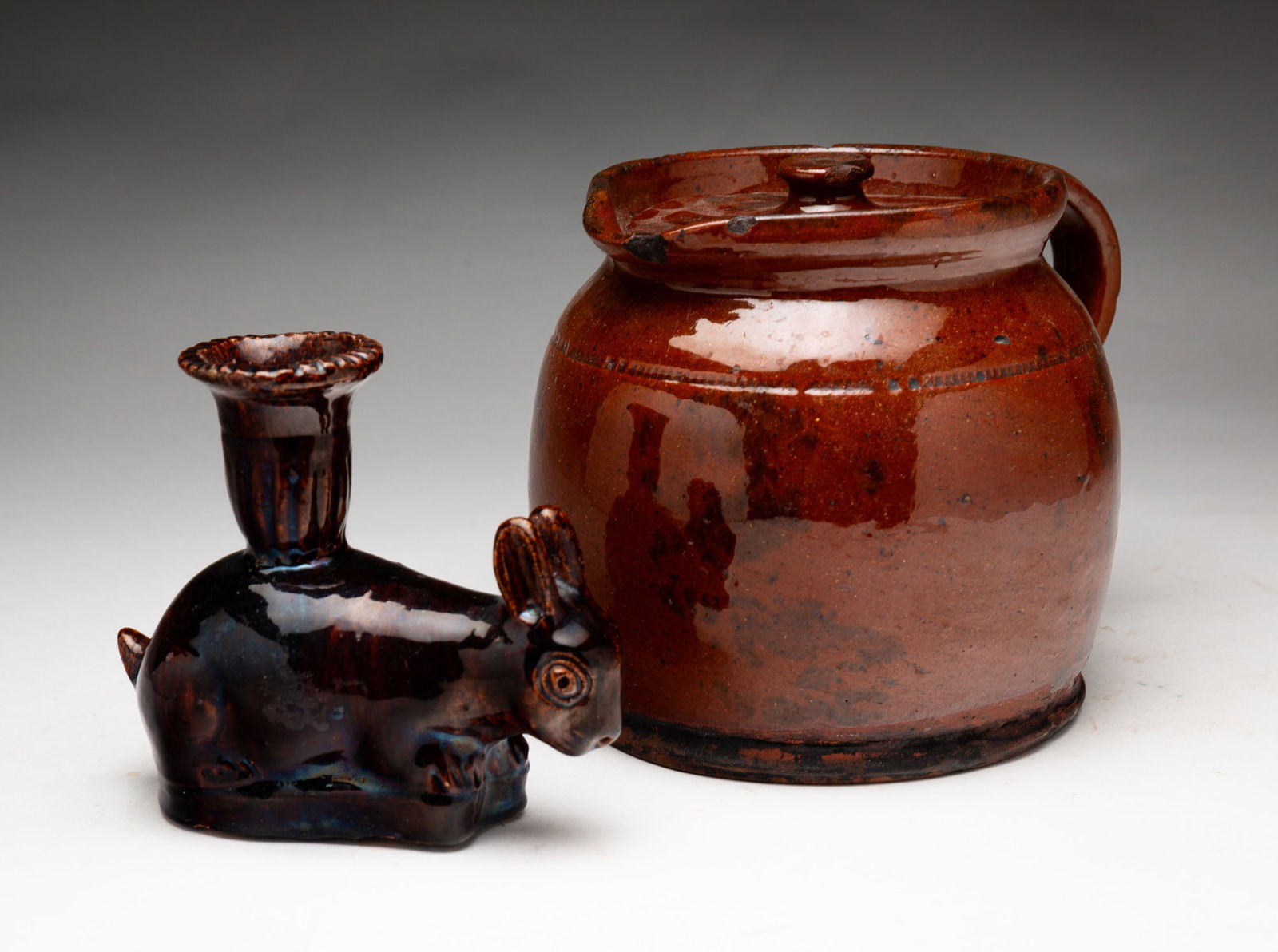 TWO PIECES OF REDWARE.: American, mid-19th century. Lidded pot with coggled decoration, edge wear and loss, 5.75"h. A rabbit figural candlestick, chips and surface wear, 4.5"h.