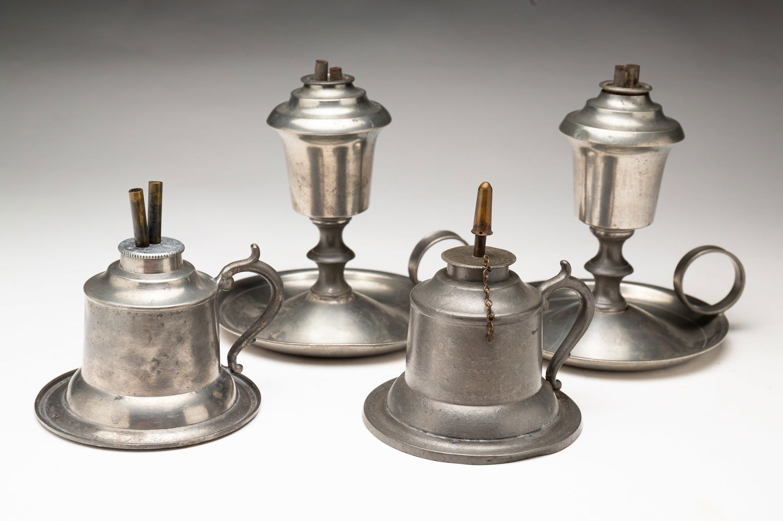 FOUR PEWTER FLUID BURNING LAMPS.: American, mid-19th Century. A pair of R. Gleason whale oil lamps with double burners, inverted acorn fonts, baluster stems and handled saucer bases, 6"h. And a near pair of Capen & Molineux bell shape