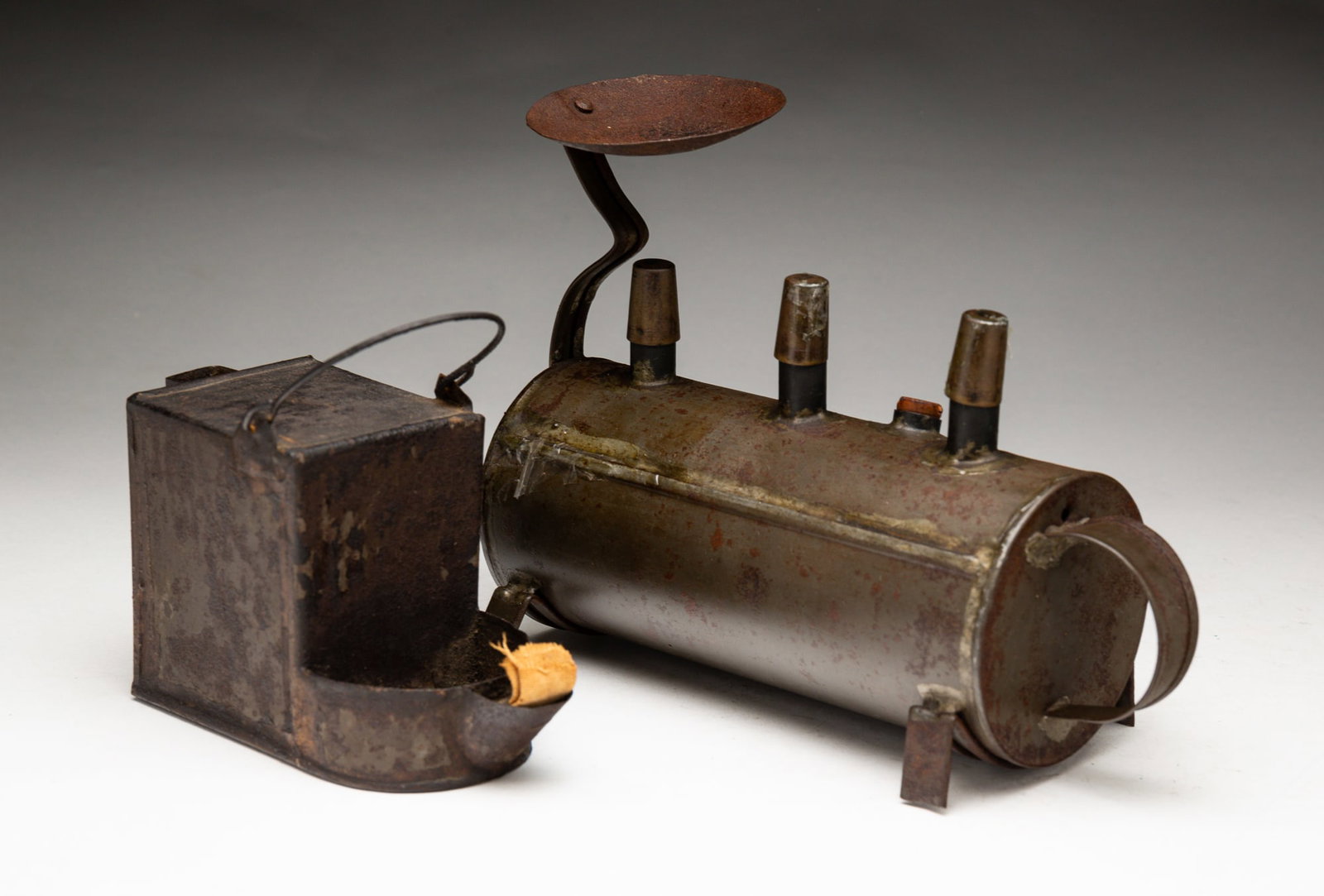 TWO TIN LAMPS.: American, second quarter 19th Century. A box shaped grease lamp with wire bale handle and worn black paint. 3.5''h. plus handle. And a three-burner whale oil lamp on a horizontal cylindrical tank, hal