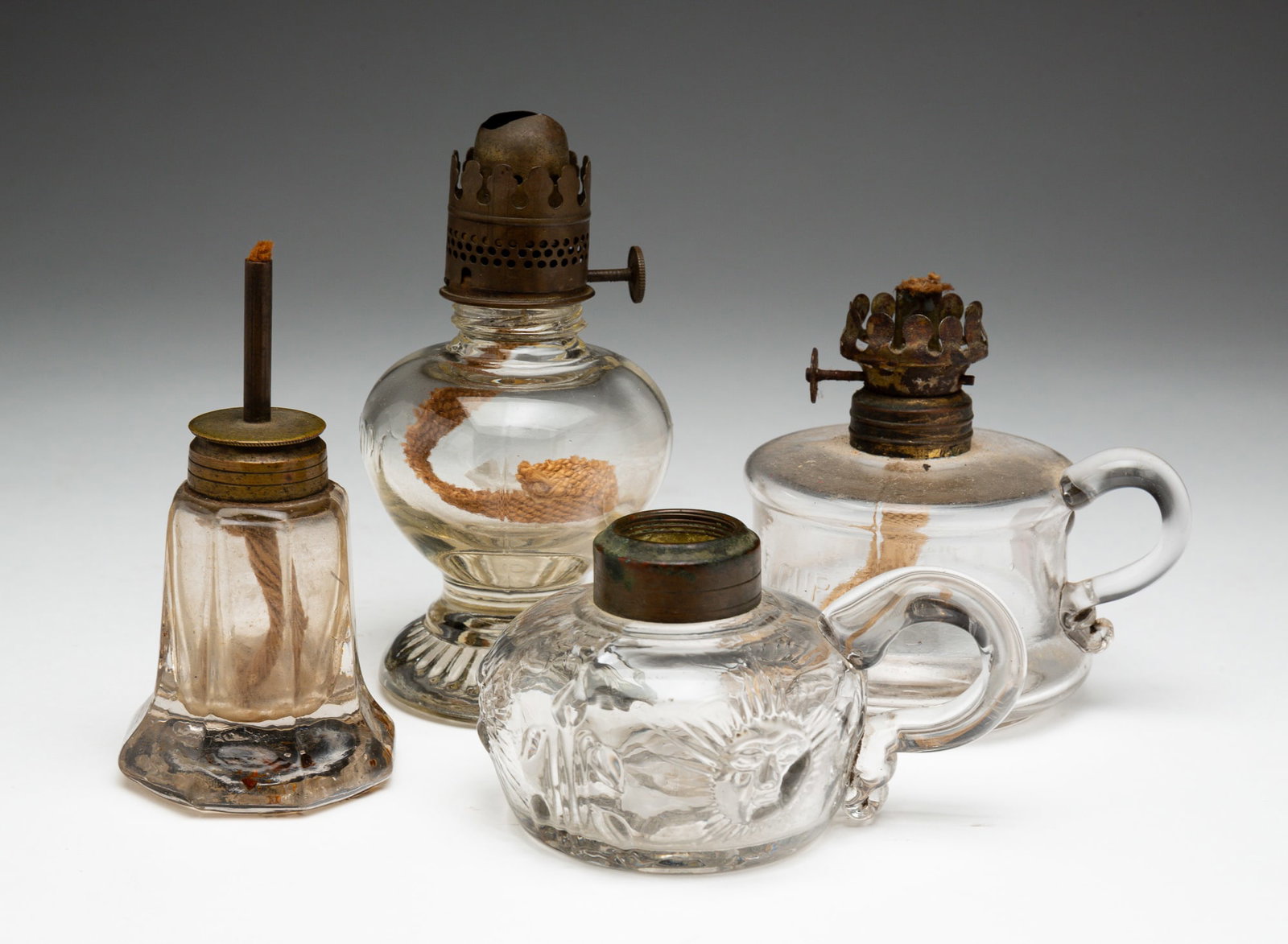 FOUR VARIOUS PRESSED GLASS SMALL LAMPS.: American, second half 19th Century. Soda lime glass. Two finger lamps: the smallest with Sunburst pattern and applied hands, 2.25"h., middle size embossed "Little Butter Cup". 3.5"h.And a footed lamp 