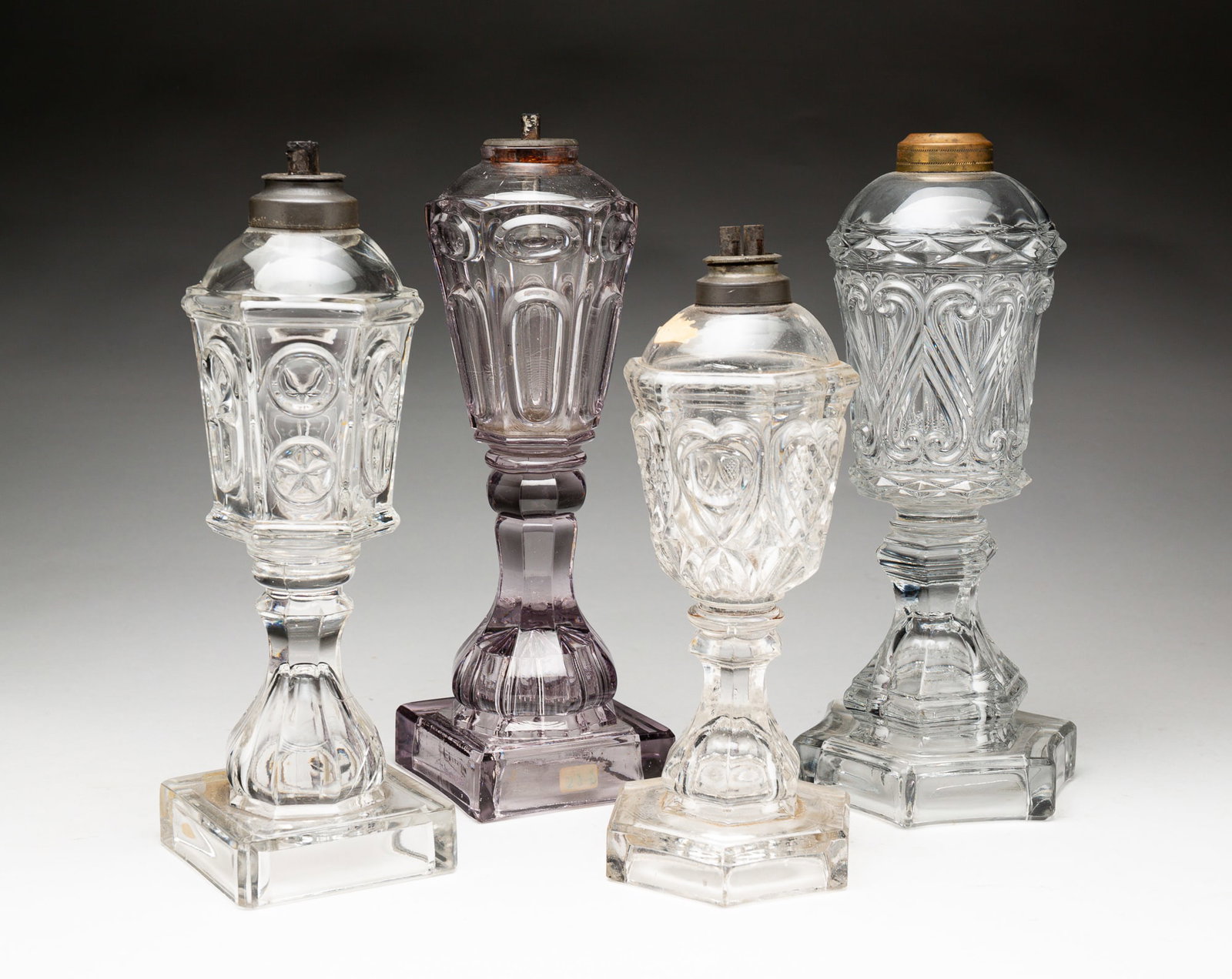 FOUR SANDWICH GLASS WHALE OIL LAMPS.: American, mid-19th Century. Flint glass. One has amethyst tinge with buckle pattern variant pattern on a square base. 10.35''h. A heart pattern variant with curved hexagonal base, 10"h. A "Star and Pr