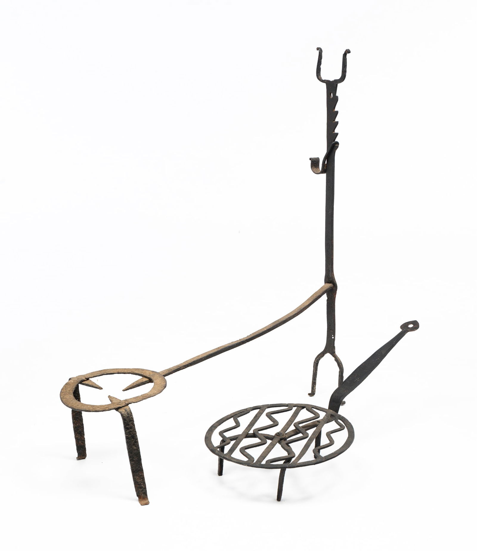 TWO WROUGHT IRON FIREPLACE TOOLS.: American, first quarter 19th Century. A rotating broiler, heart shape handle end and a zig zag pattern on table. Some wear and pitting. 8.5"h. 25.5"l. And a combination trivet, trammel and spit holder
