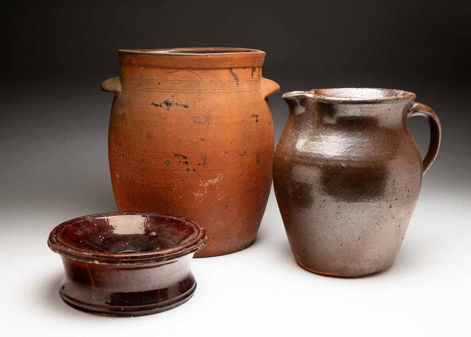 THREE REDWARE VESSELS.: American, 19th or 20th Century. Red earthenware storage jar with lug handles and glaze on interior. With scratching, chips, and crazing. 13"h. Red earthenware pitcher with brown glaze and handle and s