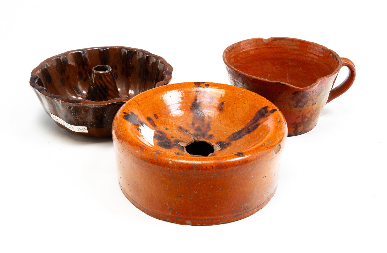 THREE PIECES OF REDWARE.: American, mid-19th Century. All three have mottled glaze decoration. A Turk's head mold. Wear and a couple small chips. 3.25"h. 9"dia. A cuspidor. Chips and glaze loss. 4"h. 9.5"dia. And a handled spo