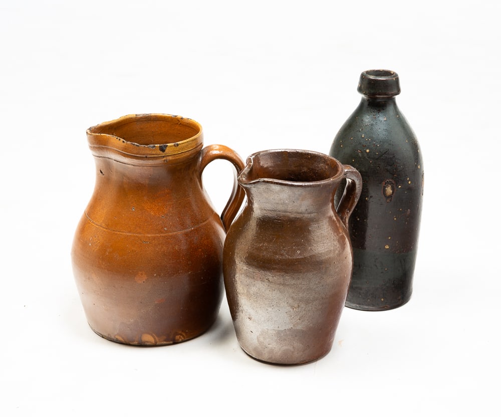 TWO SALT-GLAZED STONEWARE PITCHERS AND A BOTTLE.: American, second half 19th Century. Pitcher with brown glaze and incised band around shoulder. Several old chips throughout, especially to top rim. Heat discoloration to one side. 8"h. Redware pitcher