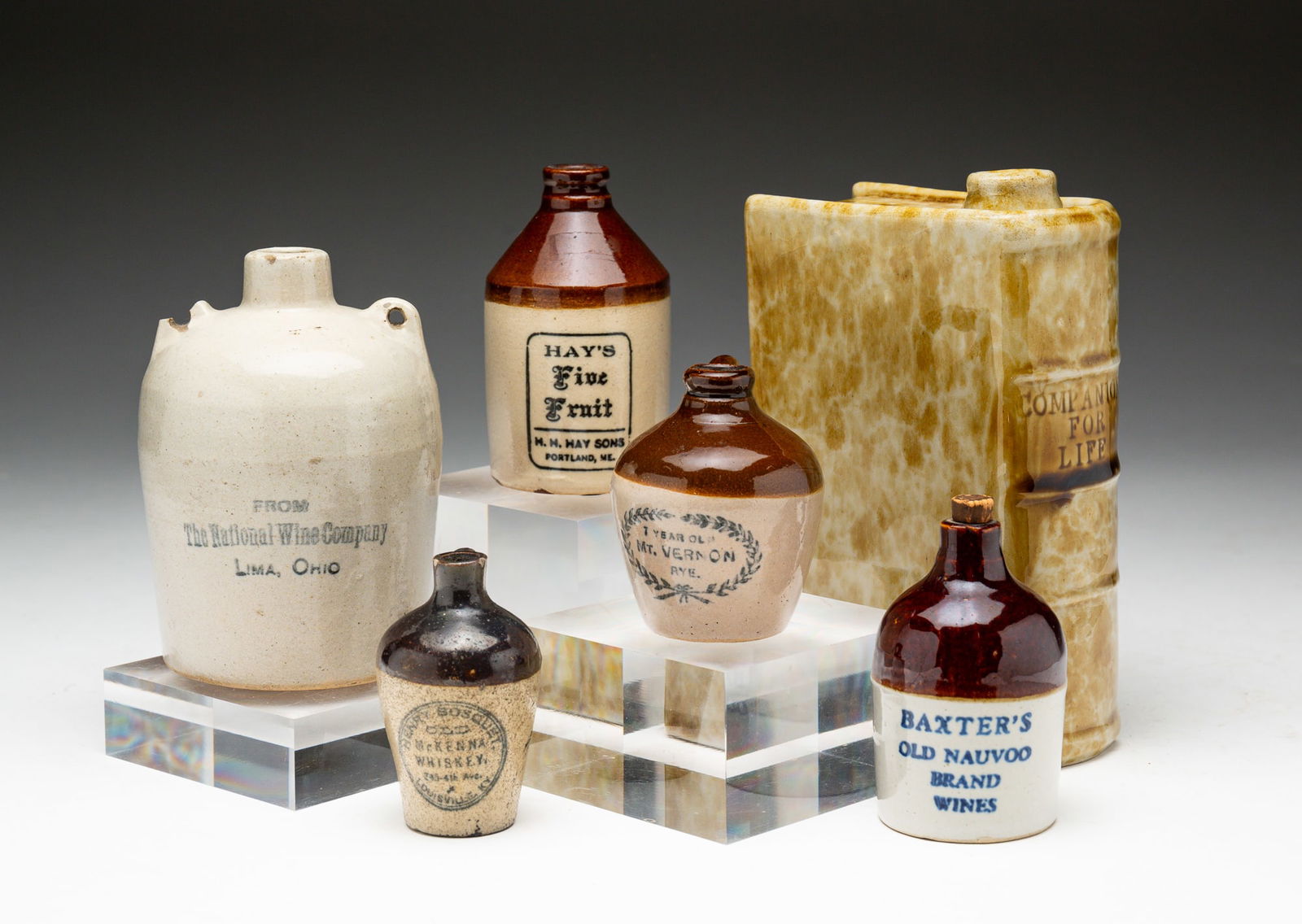 FIVE ADVERTISING MINIATURE JUGS AND A REPRODUCTION BLOCK BENNINGTON BOOK FLASK.: Jugs circa 1900, flask circa 1980. Four salt-glazed stoneware two-tone brown and cream jugs reading "BAXTER'S OLD NAUVOO BRAND WINES", "MCKENNA WHISKEY", "7 YEAR OLD MT. VERNON RYE." and "HAY'S FIVE F