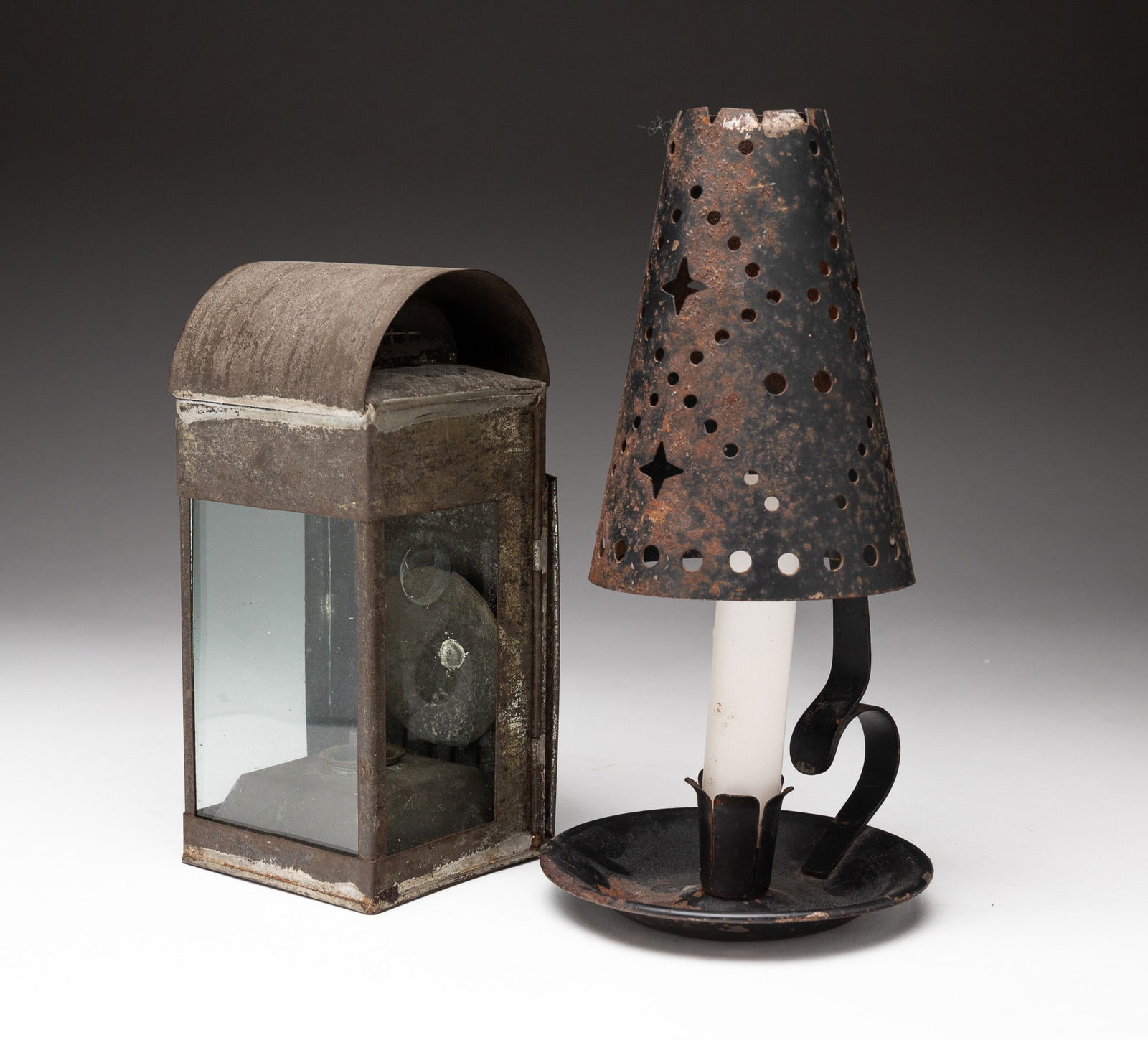 TWO LIGHTING DEVICES.: American, mid-19th and early-20th Century. Punched tin shade on candlestick, oxidation and wear, 9"h. Small tin oil lantern with folding handle and glass sides, wear and loss, 7"h.