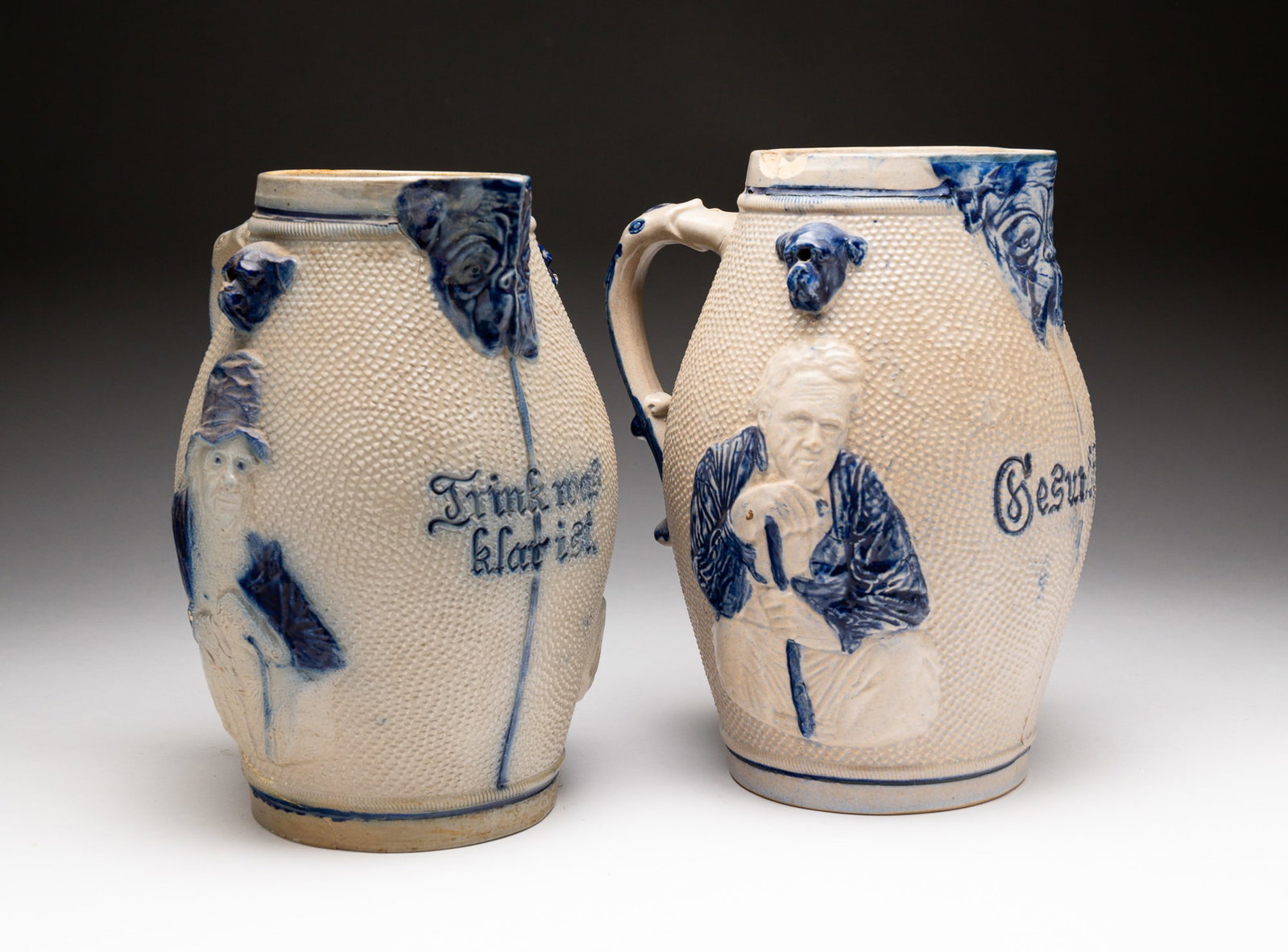 TWO BLUE AND GRAY SALT-GLAZED STONEWARE PITCHERS.: American, late-19th Century. Attributed to Whites Pottery, Utica, NY. First pitcher molded with portraits of men and embossed with "Trink was klar ist" ("Drink what is clear"). Embossed "3" on undersi