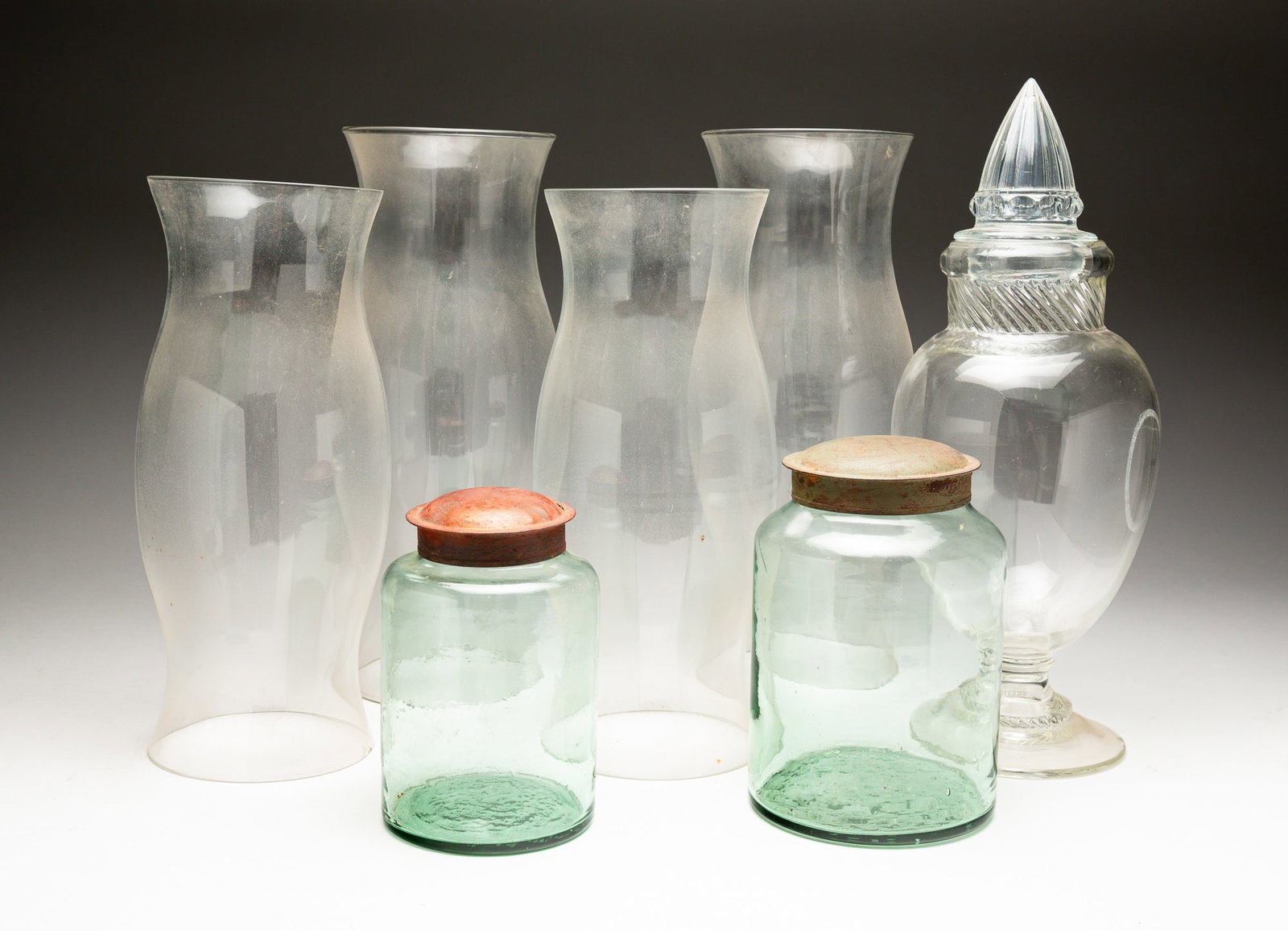 SEVEN GLASS ITEMS.: 20th Century. Two handblown green glass jars with metal lids. With chips to top rims. 7"h. And 8"h. Molded glass pedestal candy jar with mismatched lid. 16"h. Four glass hurricanes. With a chip to two
