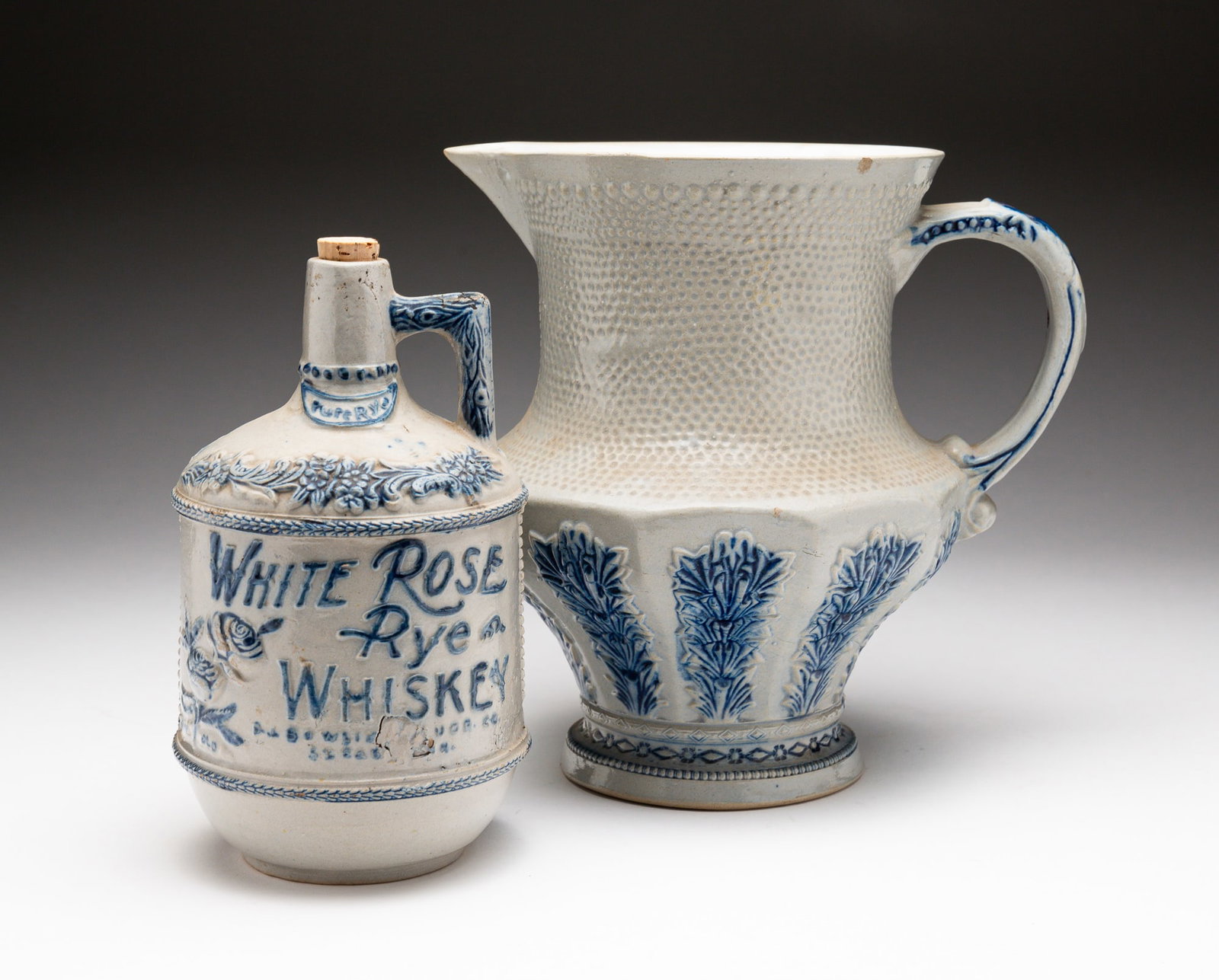 BLUE AND GRAY STONEWARE WHISKEY JUG AND PITCHER .: American, late-19th Century. The jug molded "WHITE ROSE RYE WHISKEY P.J. BOWLIN LIQUOR CO. ST. PAUL MINN." with floral decoration and molded handle. Chips, crazing, and discoloration throughout. 8"h. 