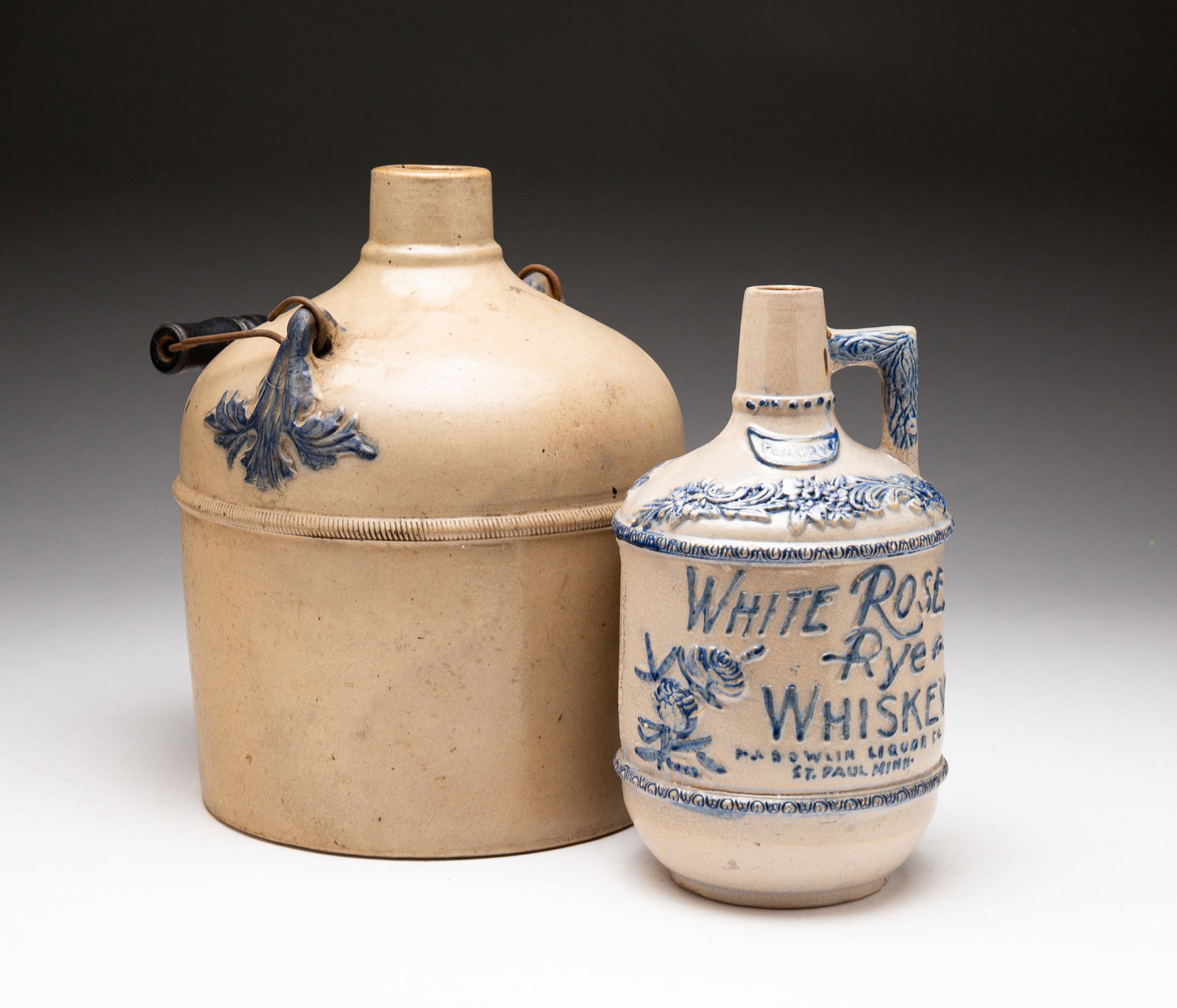 TWO BLUE AND GRAY STONEWARE JUGS.: American, late-19th Century. The first molded "WHITE ROSE RYE WHISKEY P.J. BOWLIN LIQUOR CO. ST. PAUL MINN." with floral decoration and molded handle. Chips and crazing throughout. 8"h. The second jug