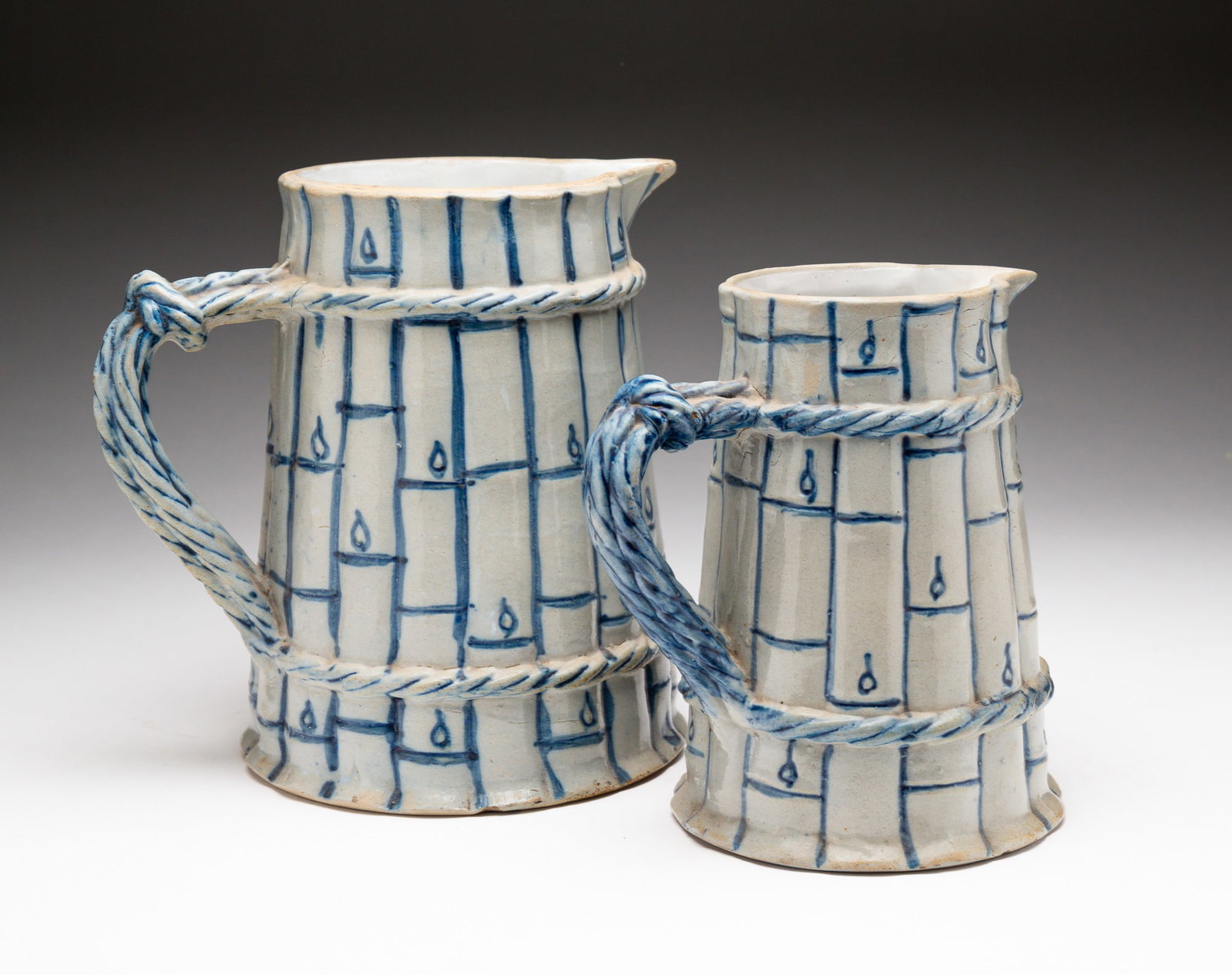 TWO BLUE AND GRAY STONEWARE PITCHERS IN BAMBOO MOTIF.: American, late-19th Century. Attributed to Whites Pottery, Utica, NY. Each molded in bamboo motif with rope motif handle. The larger embossed with "4" on underside. Chips to top and bottom rims with h