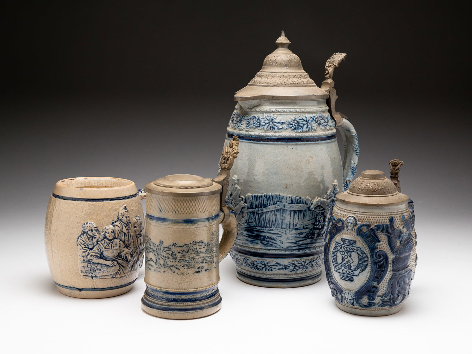FOUR BLUE AND GRAY SALT-GLAZED STONEWARE VESSELS.: American, late-19th Century. Attributed to Whites Pottery, Utica, NY. Consisting of three steins and one mug. The first stein molded with figures and flora and a medallion embossed with overlapping Gr