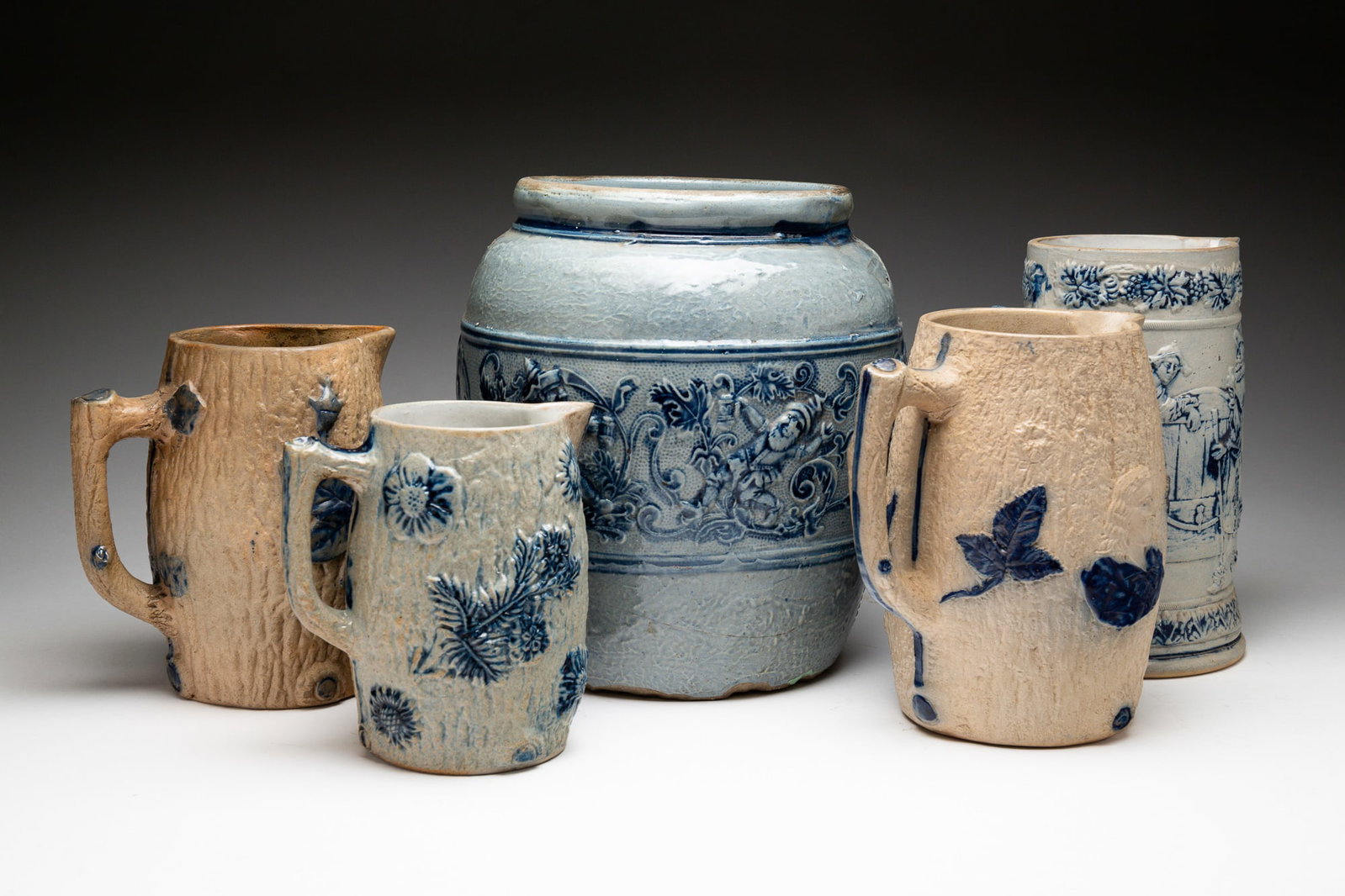 FIVE BLUE AND GRAY SALT-GLAZED STONEWARE VESSELS.: American, late-19th Century. Comprising three molded pitchers and a molded stein. The stein molded with tavern scenes and embossed "5" on underside; a pitcher molded with flowers and a seated figure e