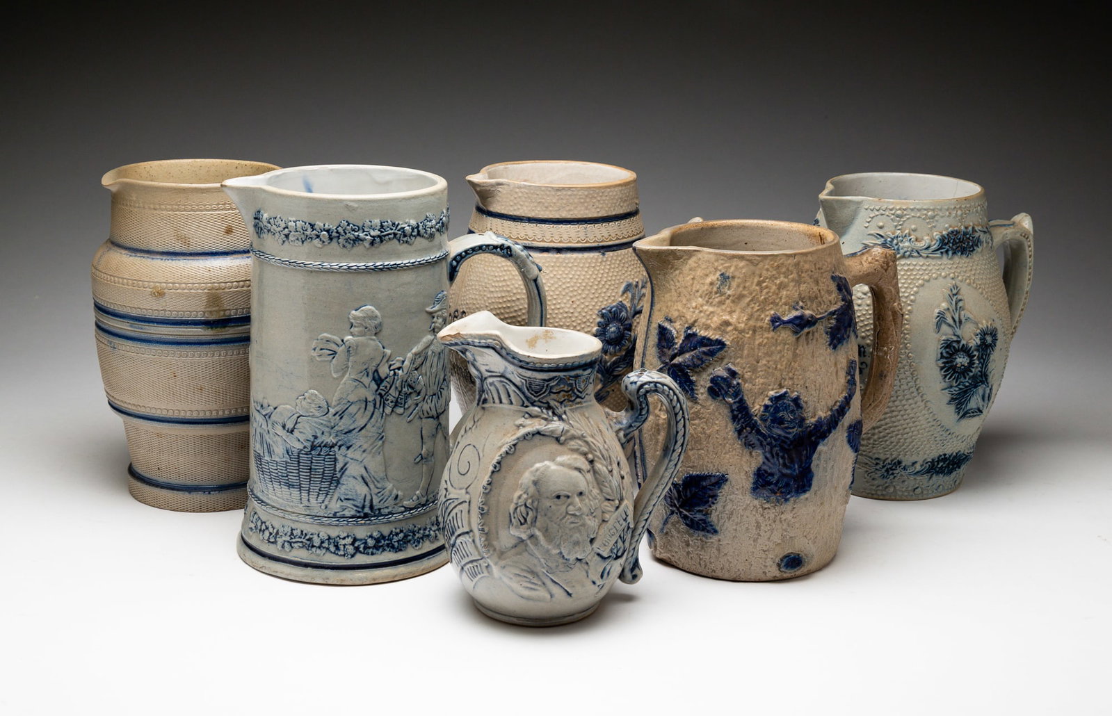 SIX BLUE AND GRAY SALT-GLAZED STONEWARE VESSELS.: American, late-19th Century. Attributed to Whites Pottery, Utica, NY. Comprising five pitchers and one stein. Stein molded with scenes of figures and embossed with "5" on underside; the smallest pitch
